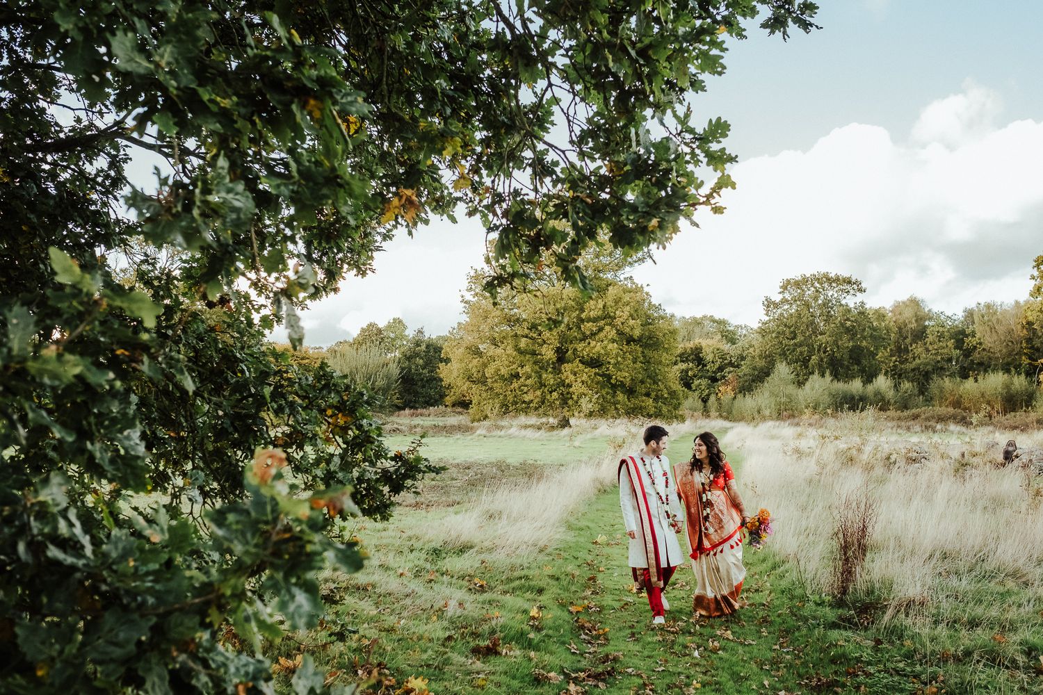 Newlyweds in traditional Indian wedding outfits walking through a serene meadow.