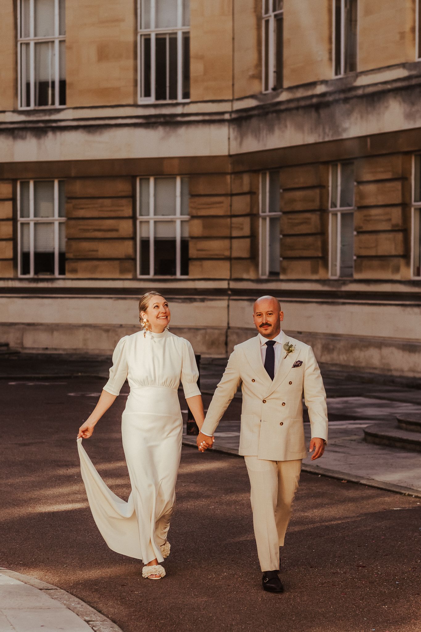 A couple in elegant all-white outfits walk hand in hand on a city street near a historic stone building in warm sunlight.