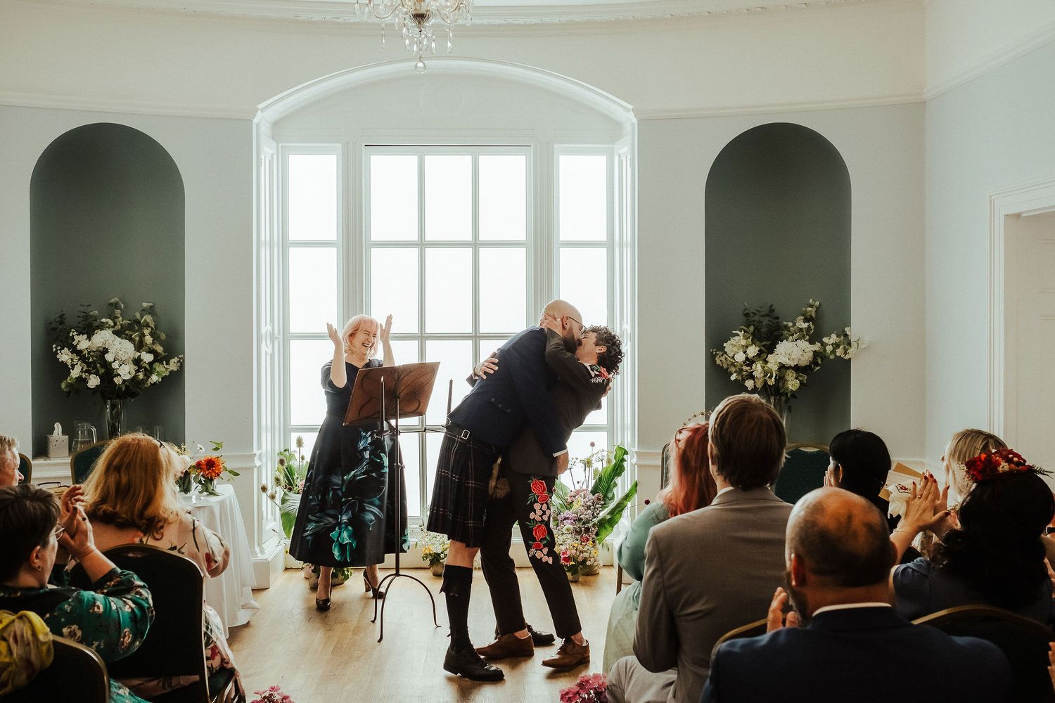 Wedding ceremony in a bright arched room as a couple kisses at the front while a celebrant and seated guests look on.