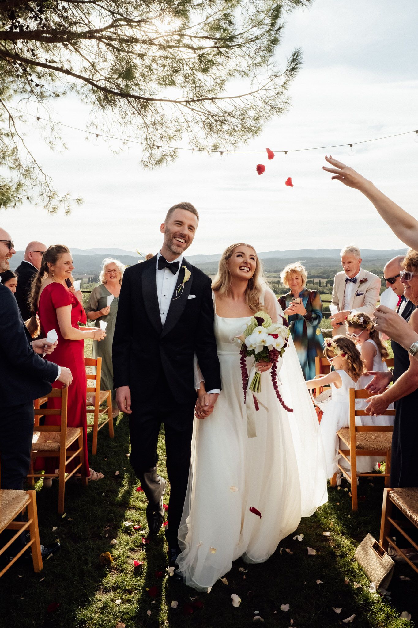 Smiling bride and groom walk hand in hand down an outdoor aisle as guests toss rose petals at a hilltop wedding ceremony.