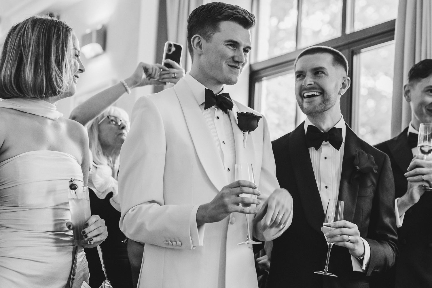 Wedding guests in formal attire gather together to take selfies and photos during a celebration.