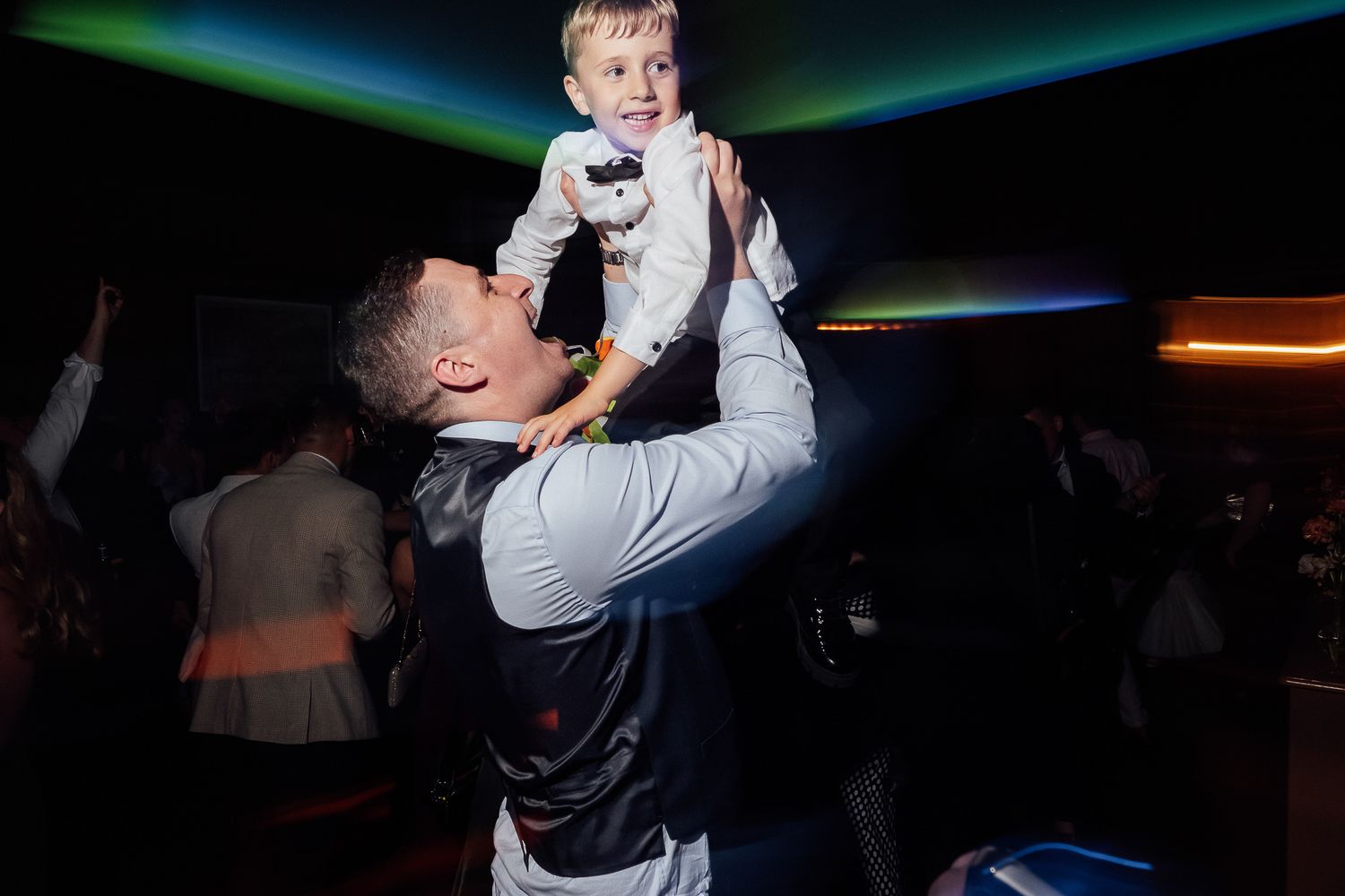 Man joyfully lifts smiling toddler above his head on a colorful dance floor at a party.