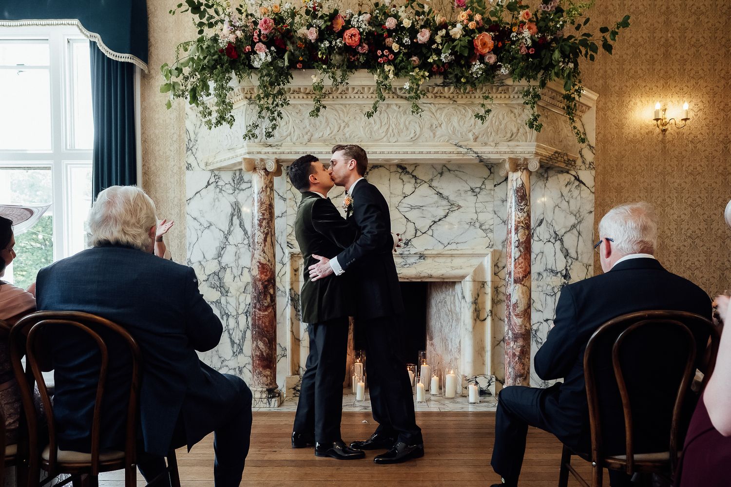 Two grooms share first kiss at elegant wedding ceremony before marble fireplace adorned with lush floral arrangement.