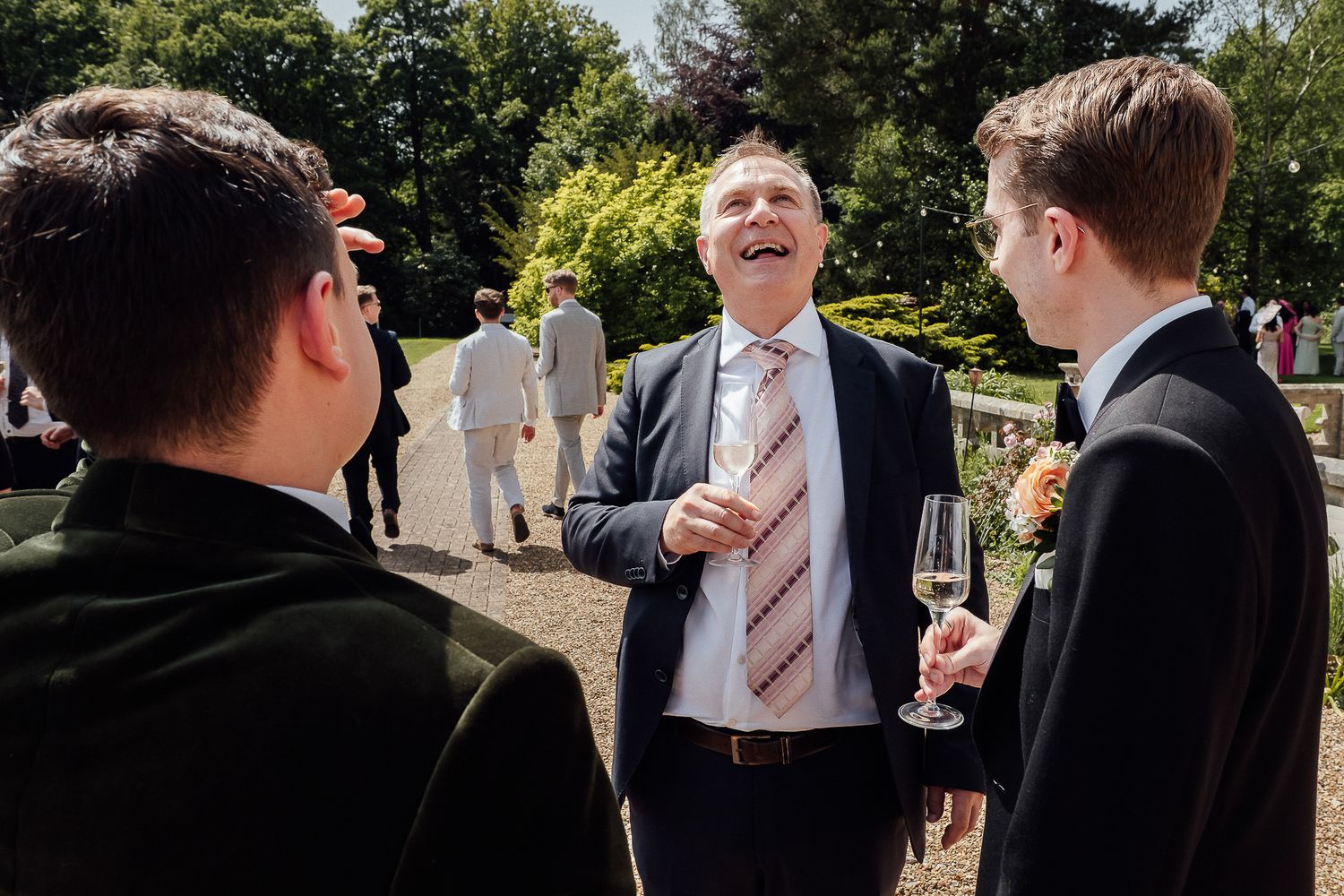 Man in suit laughing with champagne glass at outdoor wedding reception with guests in background.