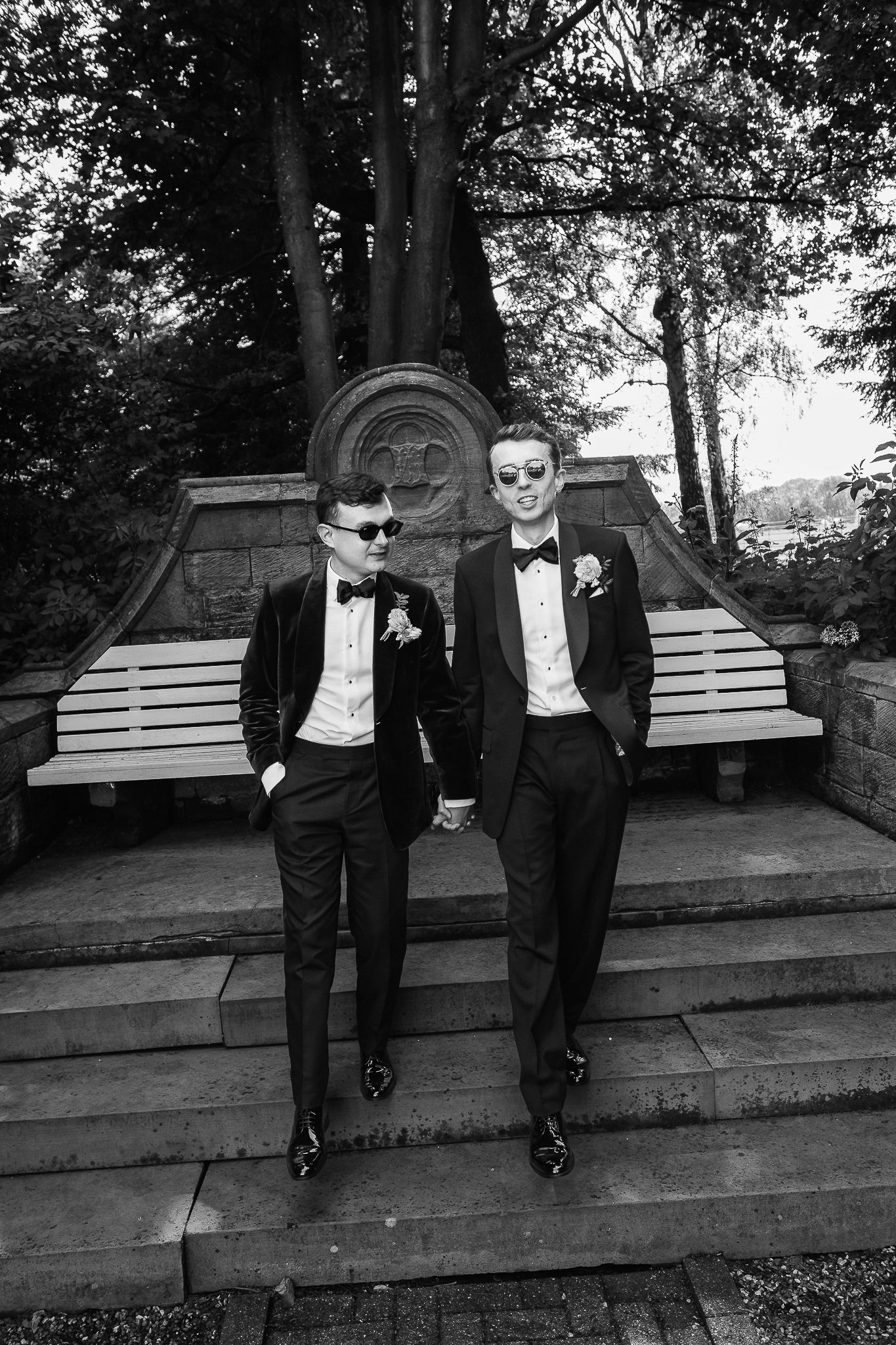 Two grooms in black tuxedos and sunglasses hold hands on stone steps in a garden wedding photo.