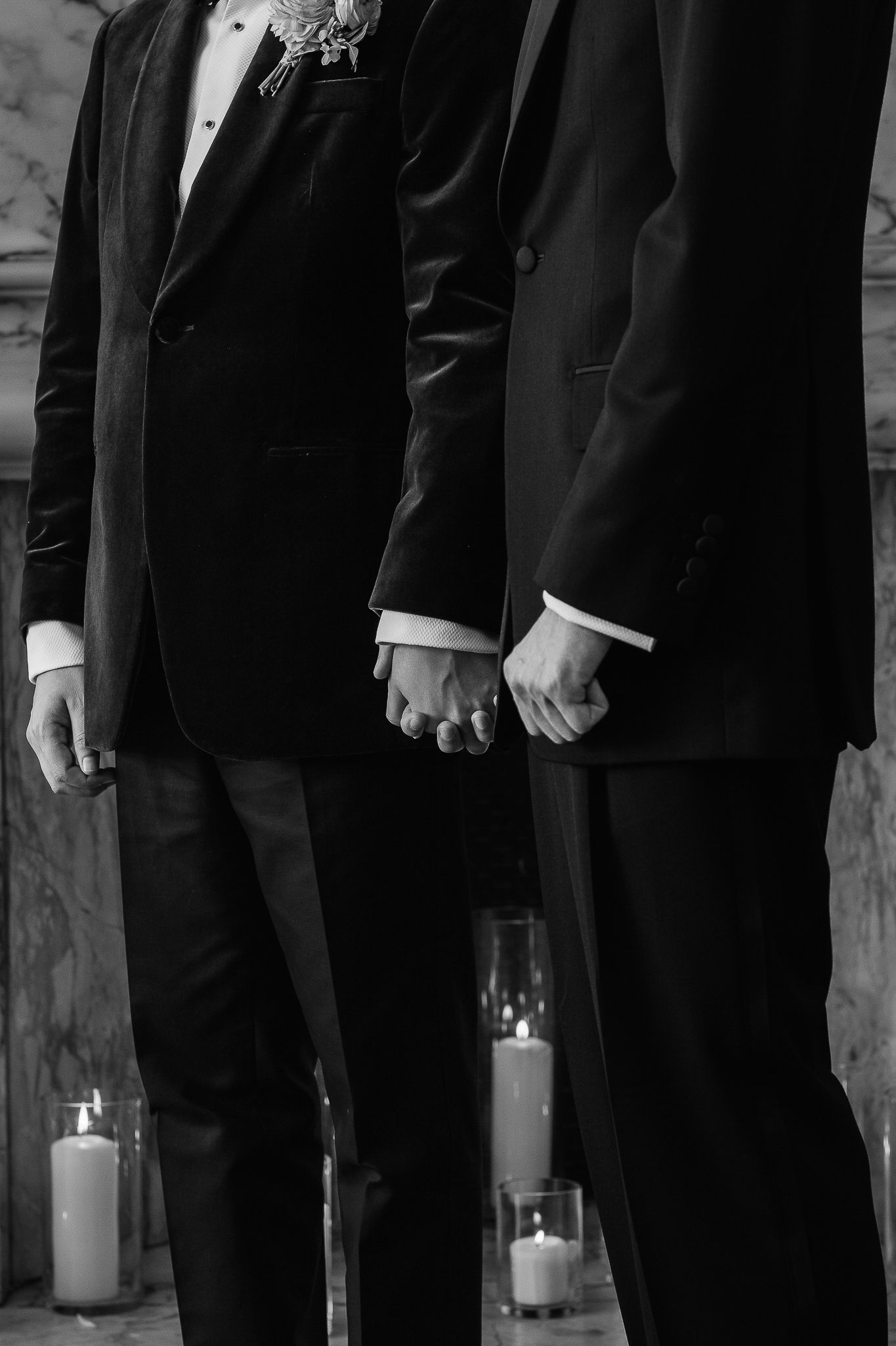 Black and white photo of two grooms in suits holding hands at wedding ceremony with candles in background.