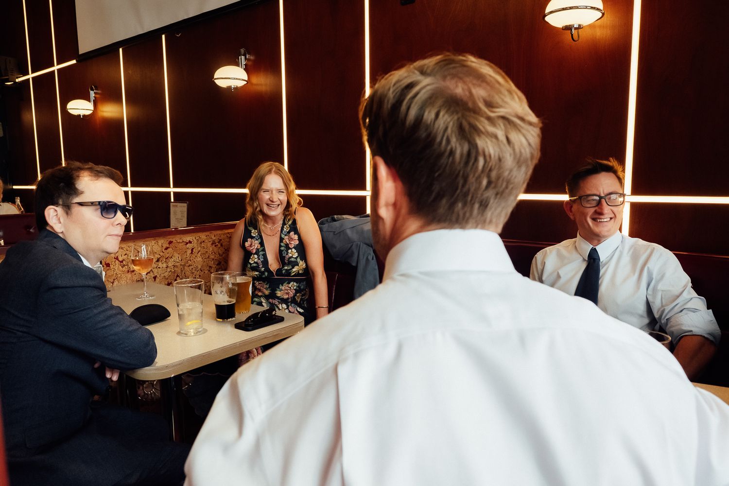 Three people chat at a cozy restaurant booth with drinks, while a man in a white shirt faces away from the camera in the foreground.