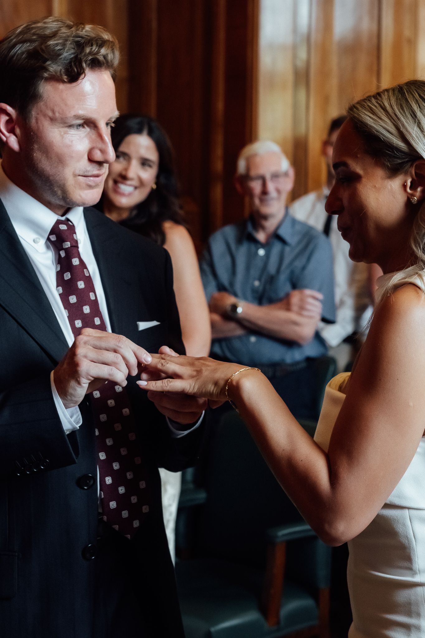 A groom in a dark suit and burgundy tie places a ring on his bride's finger during a wedding ceremony as guests smile behind them.