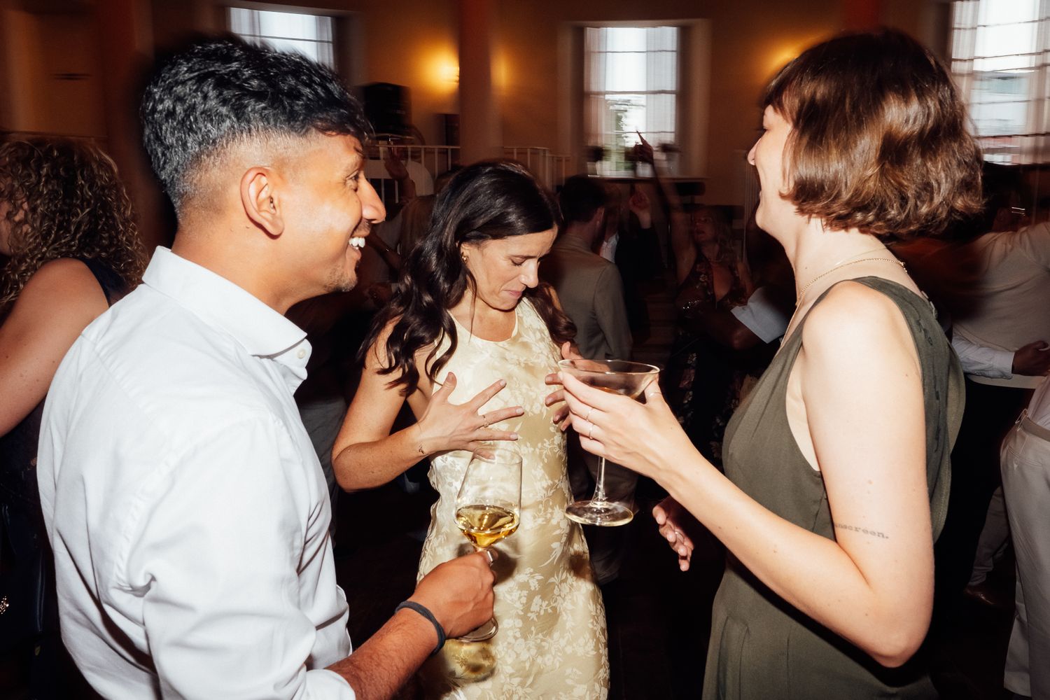 Three guests laugh and chat while holding champagne glasses at a lively indoor party with warm lighting and a crowd in the background.