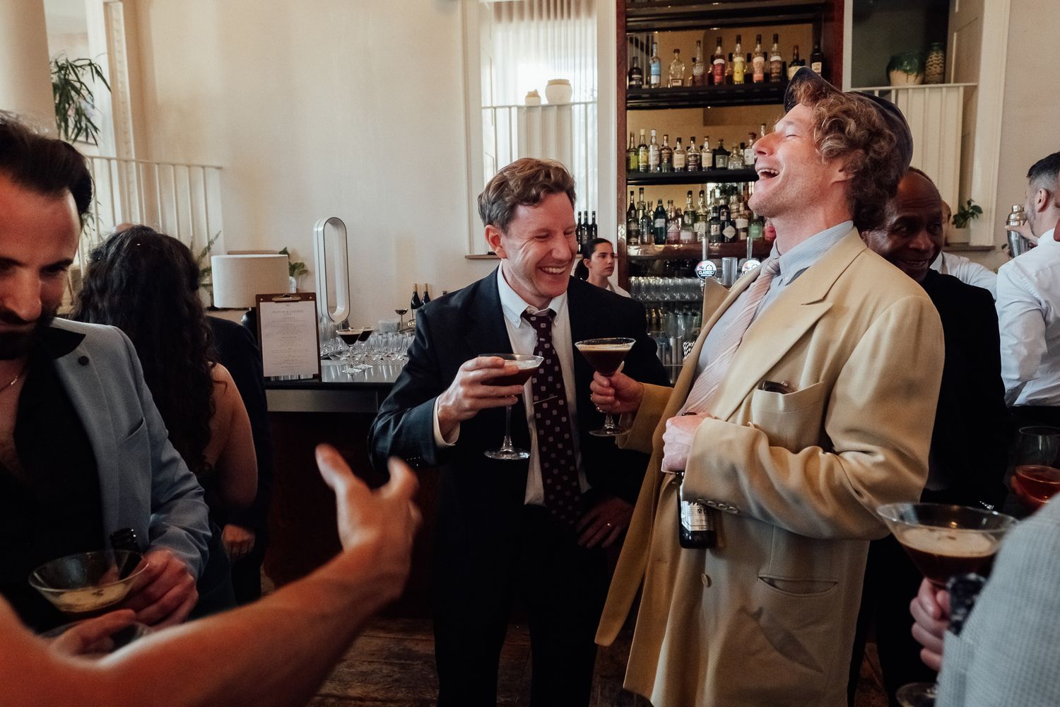Two smiling men in suits laugh while holding drinks at a lively indoor party, with a stocked bar and other guests in the background.