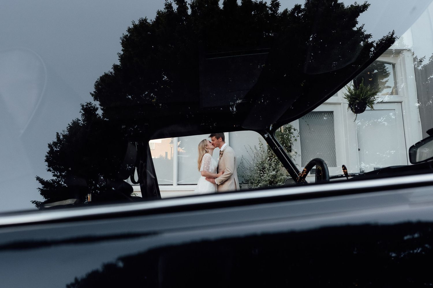 Couple embracing in a romantic moment, seen through the open hood of a black classic car with trees and a white building behind them.