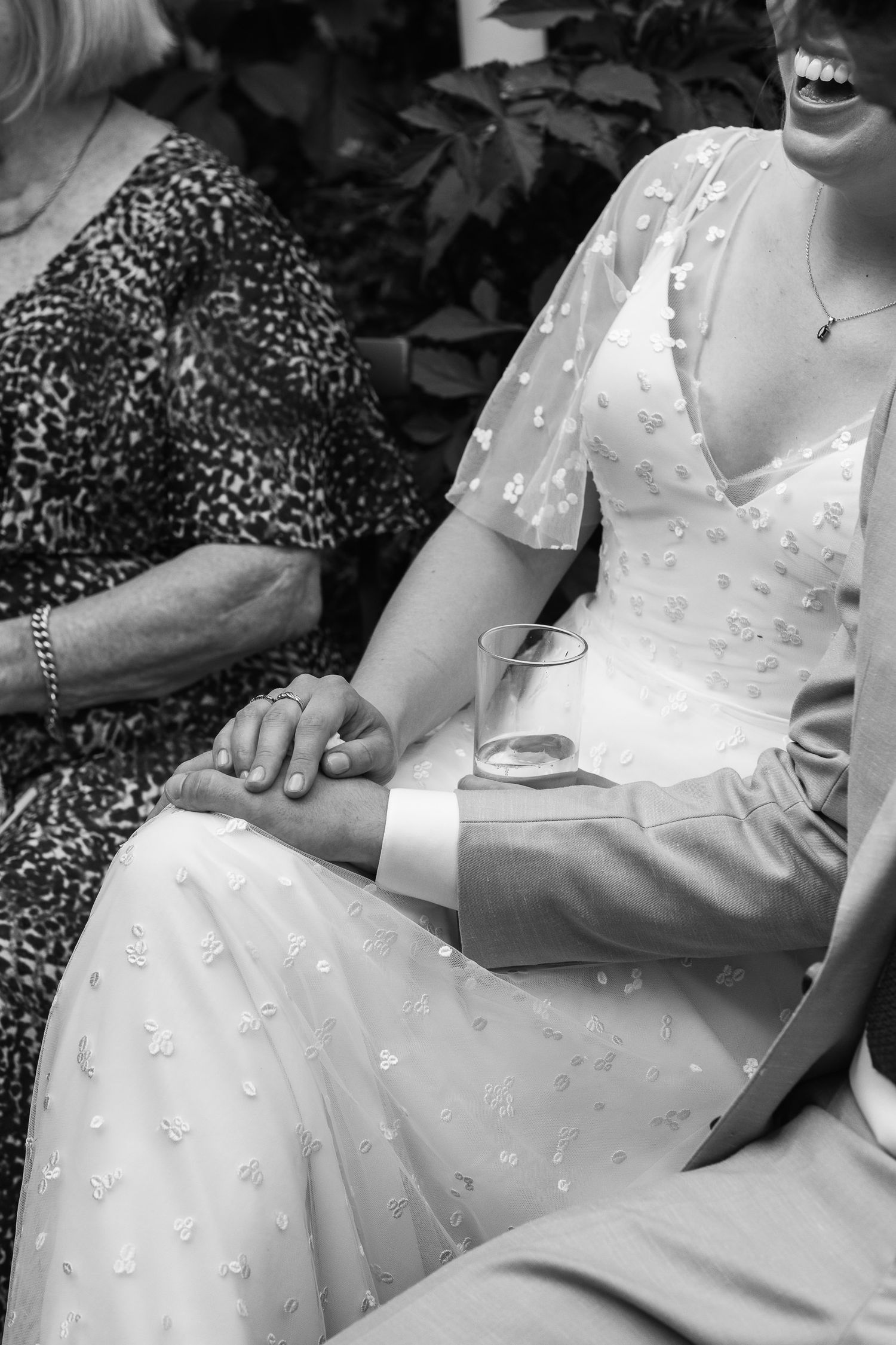 Black and white photo of a bride in a floral-embellished V-neck gown holding hands with a groom in a suit beside a smiling older woman.