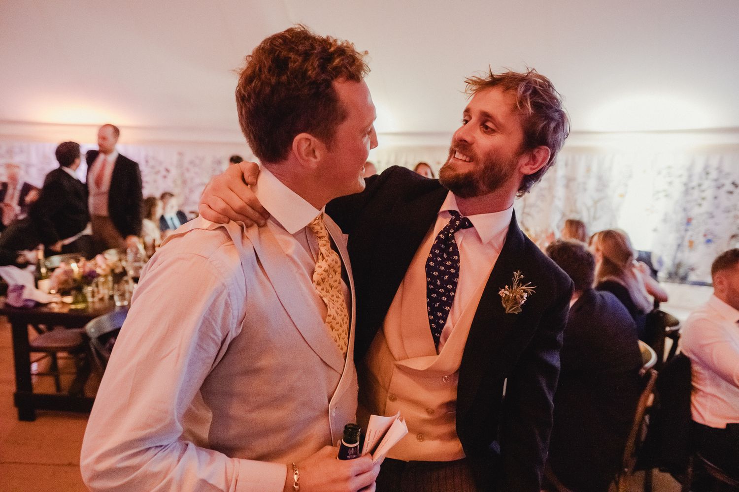 Two wedding guests in formal attire share an animated conversation at an indoor reception.
