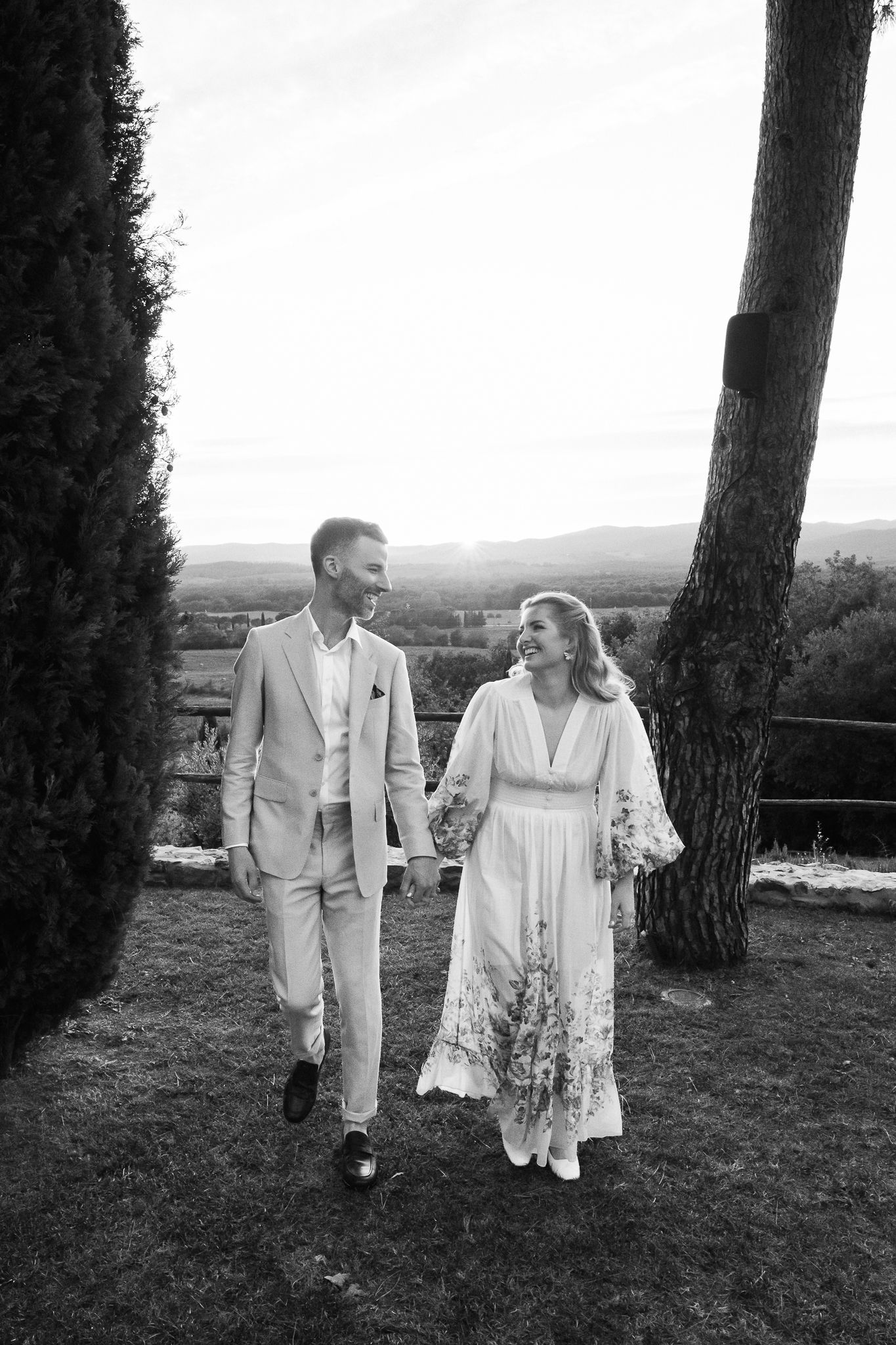 Black and white photo of a smiling couple in light wedding attire walking hand in hand on a grassy hillside overlooking distant hills.