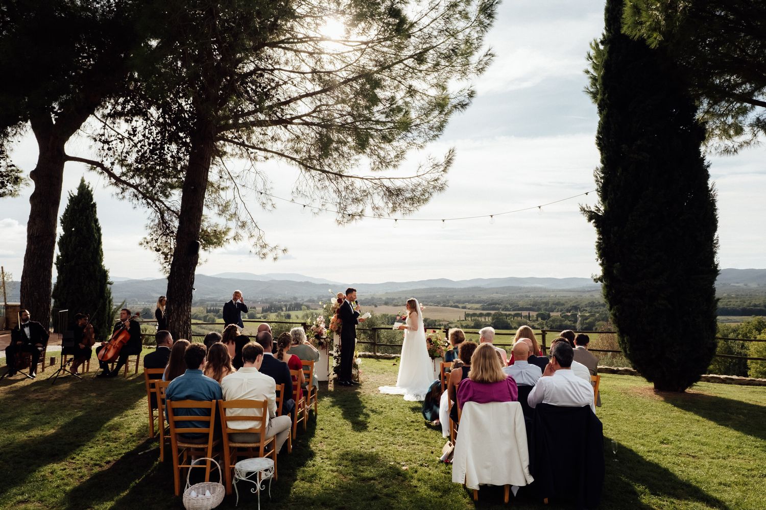 Bride in a white gown stands before seated wedding guests during an outdoor ceremony under tall cypress trees with rolling hills behind.