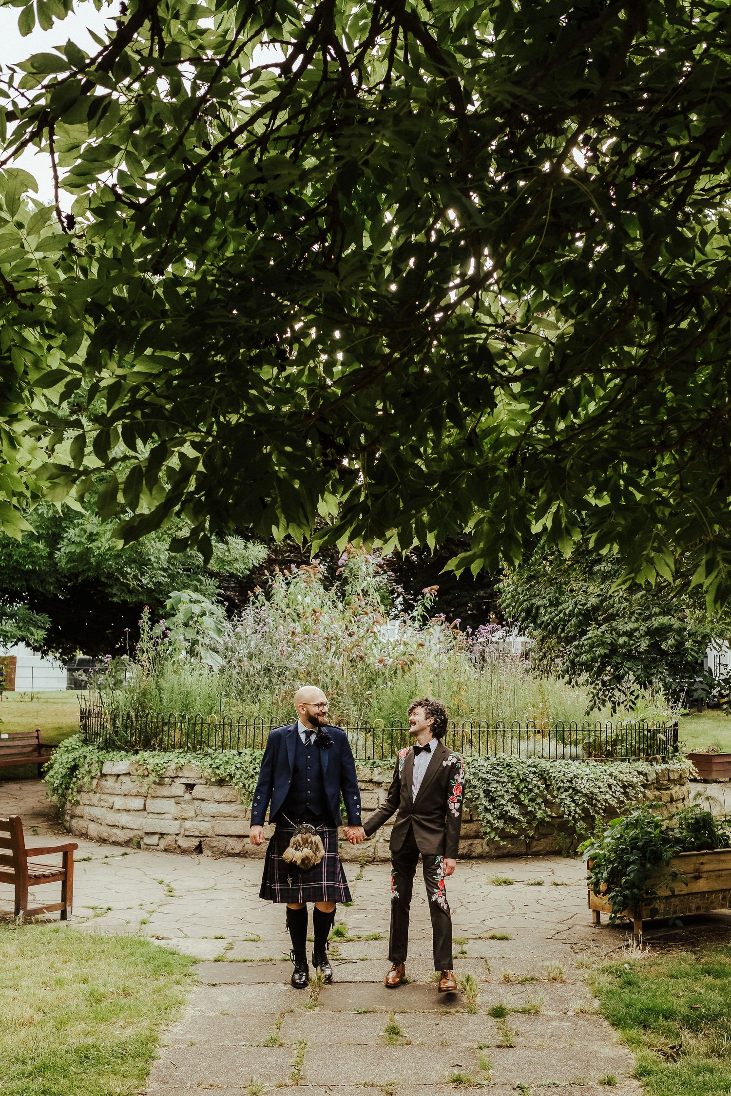 Two grooms holding hands walk along a stone garden path beneath leafy trees, one wearing a tartan kilt, at an outdoor wedding.