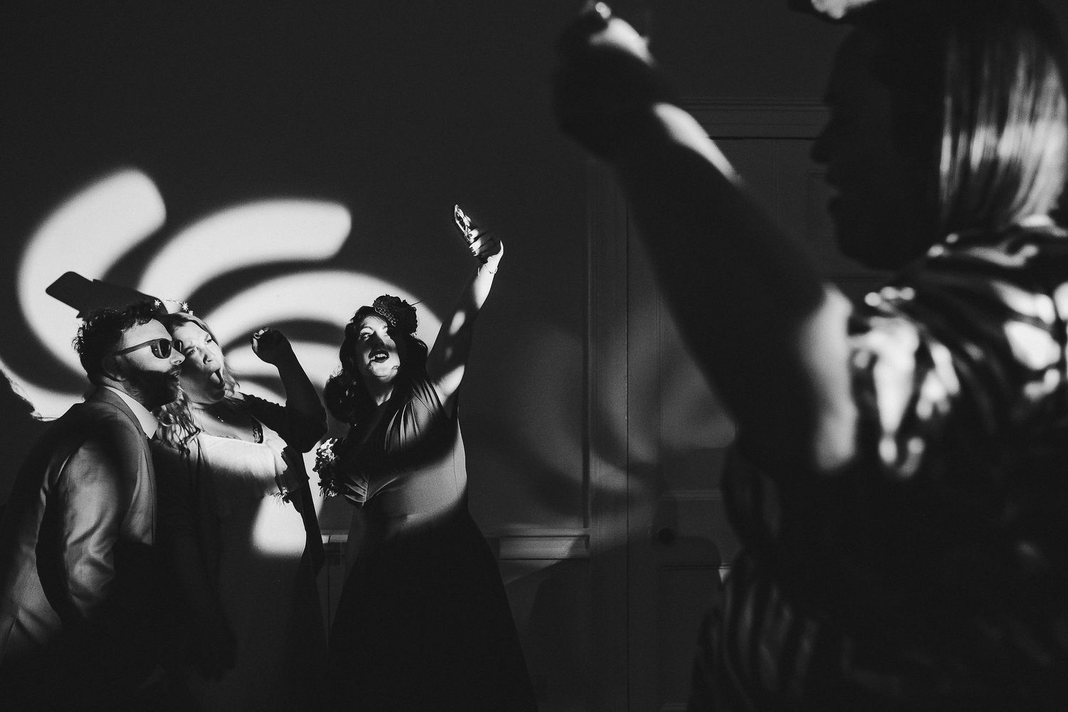 Black and white photo of guests dancing at a party with arms raised as swirling light patterns are projected on the wall behind them.