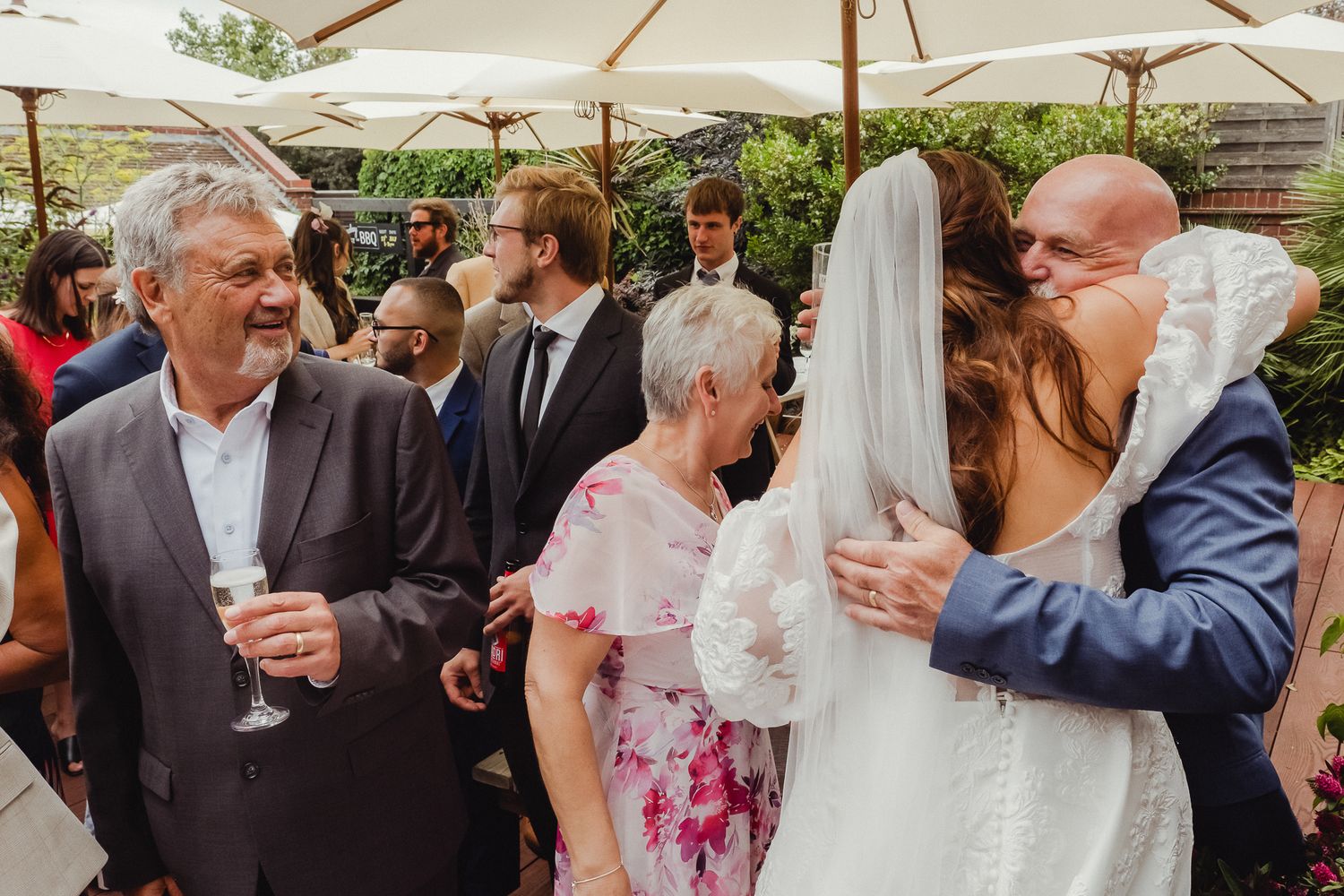 Wedding guests enjoy outdoor reception under white umbrellas with drinks and conversation in a garden setting.
