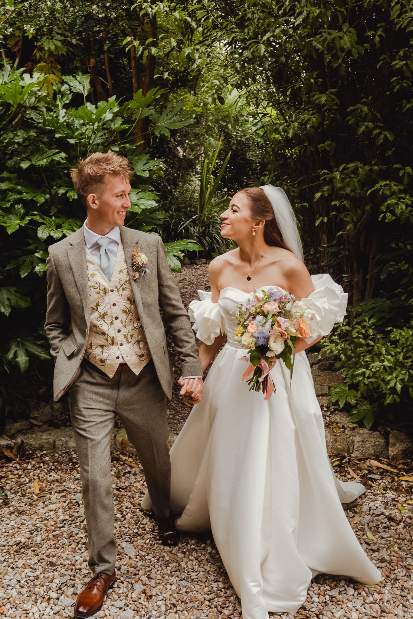A couple in wedding attire share joyful moments in a lush outdoor garden setting with natural stone elements.