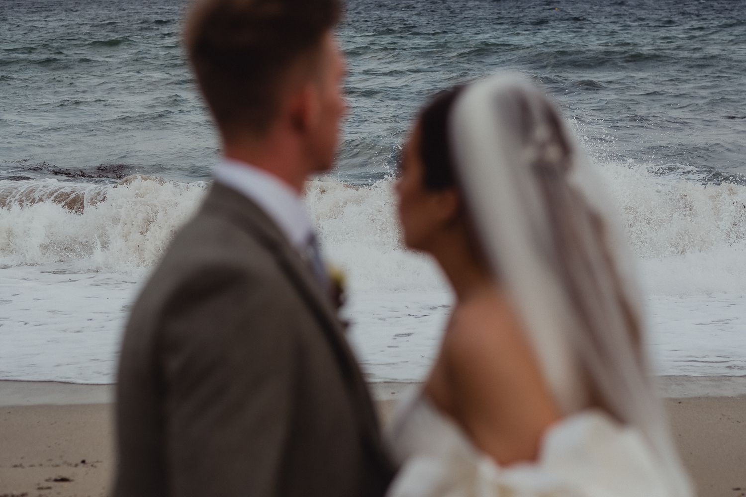 A couple in wedding attire stands together on a beach with crashing waves in the background at sunset.