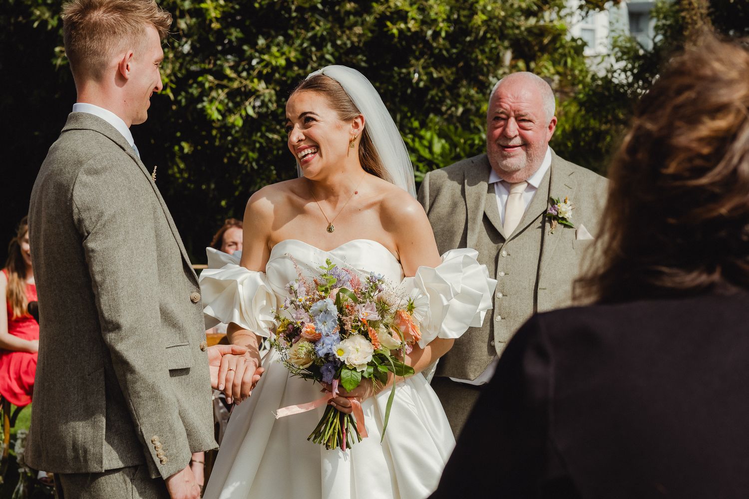 A joyful outdoor wedding ceremony moment with a bride in a white strapless gown holding a pastel bouquet.
