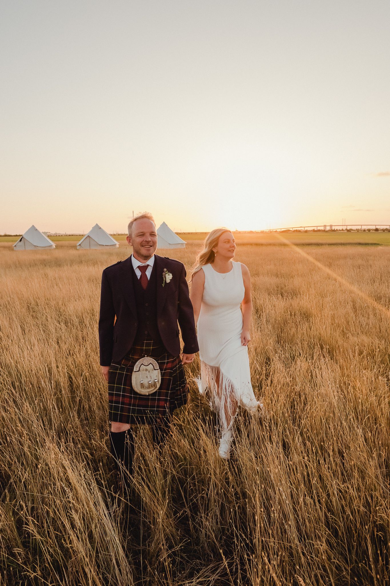 A couple walks through golden prairie grass at sunset, with the groom wearing a traditional Scottish kilt.