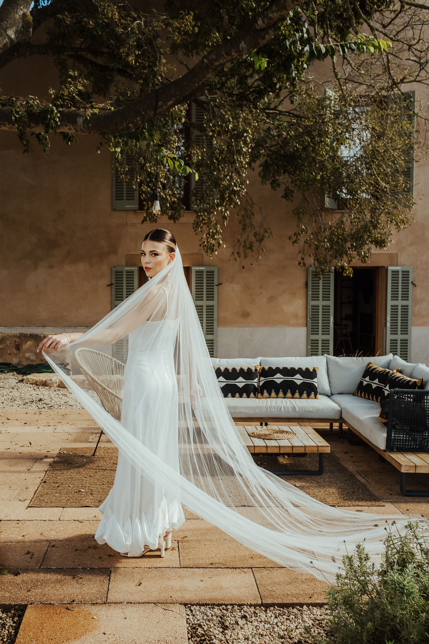 A flowing white wedding dress and veil create an ethereal scene against a rustic stone building exterior.