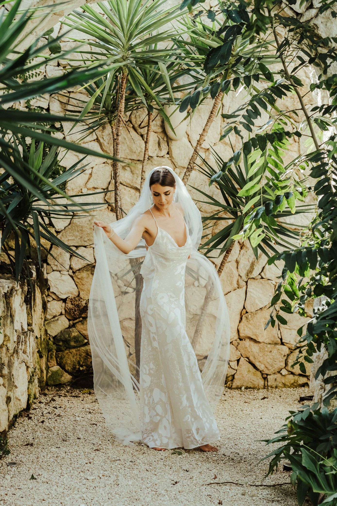 A bride in a white lace wedding dress poses gracefully in a tropical garden setting with stone walls and palm plants.