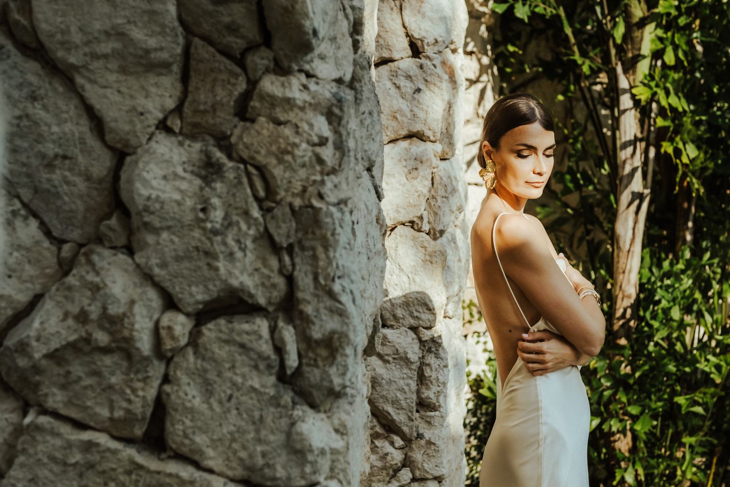 Elegant bride in white dress poses against rustic stone wall with natural greenery in outdoor setting.