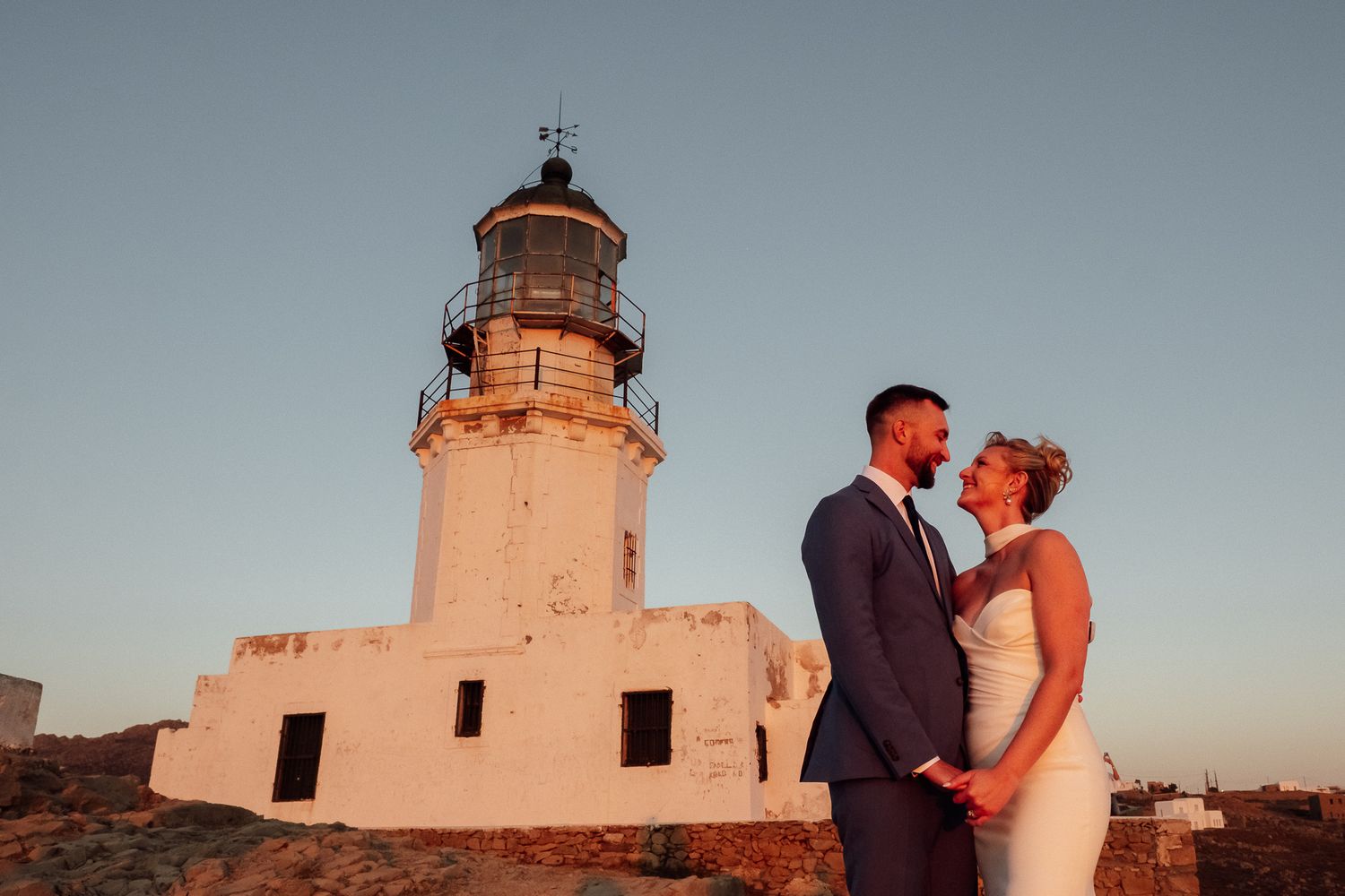 Bride and groom share a romantic moment at a white lighthouse during golden hour sunset.
