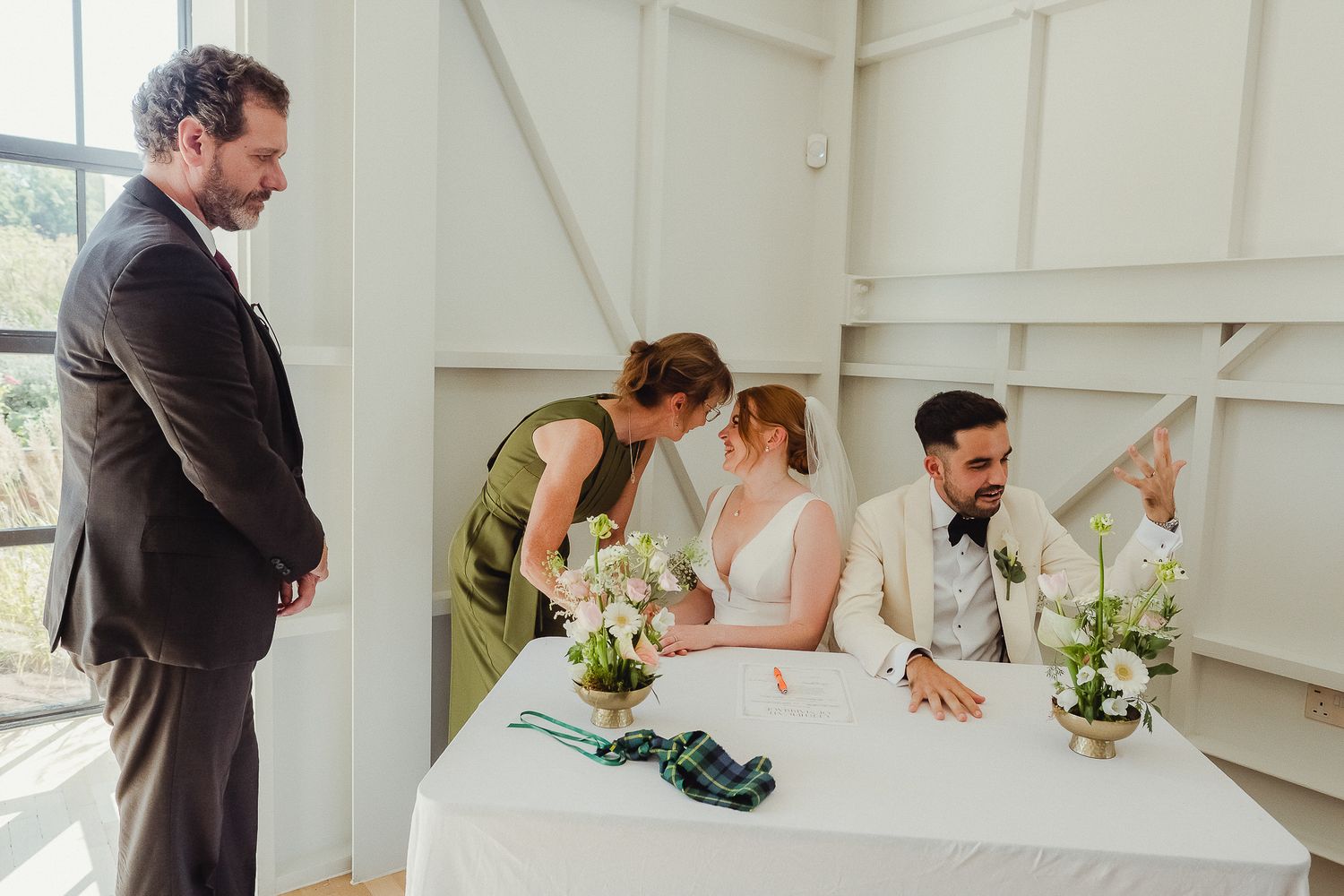 Wedding party members sign official documents during an intimate ceremony at a white-walled venue.