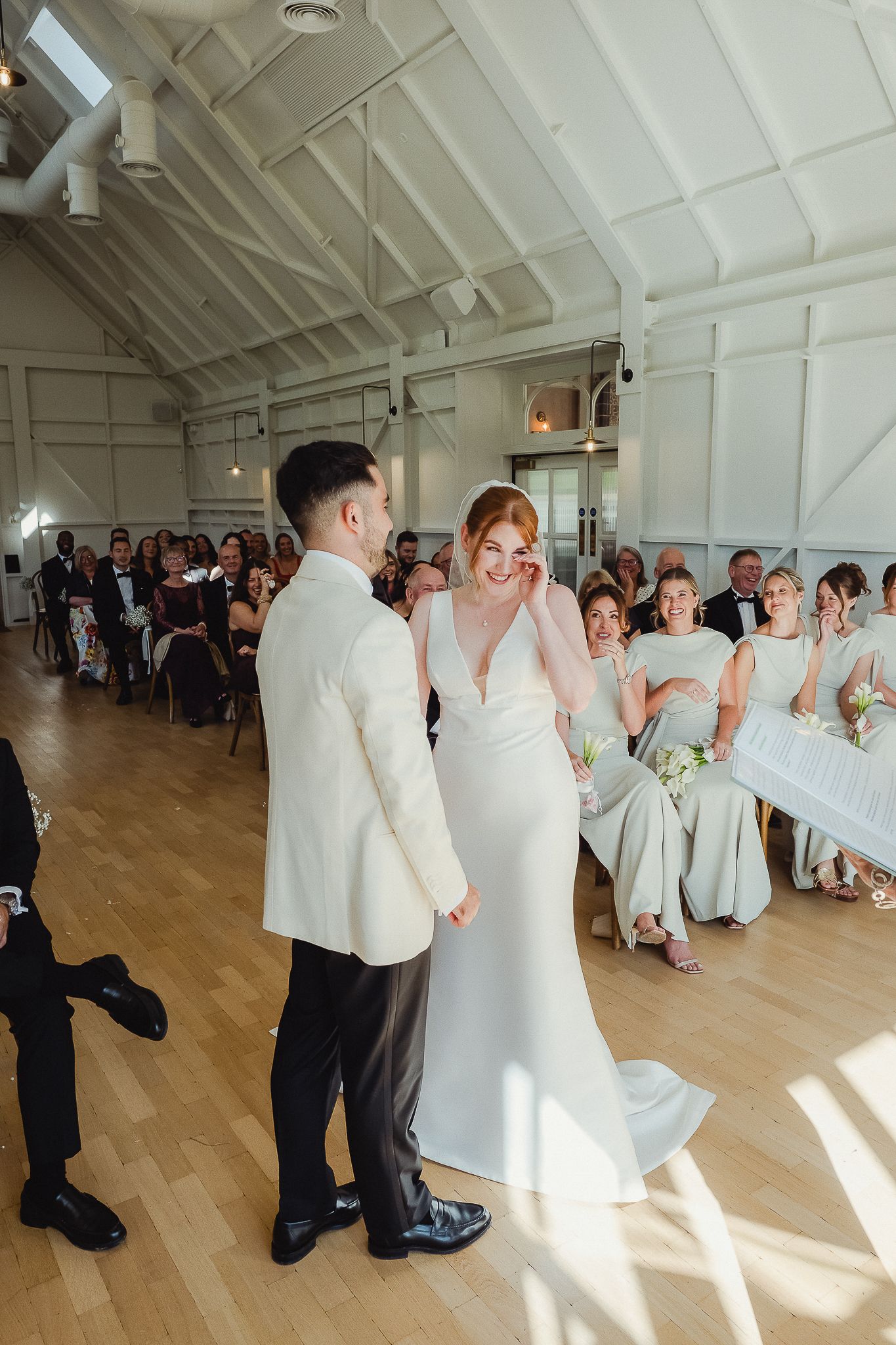 A couple shares a joyful moment during their wedding ceremony in a bright, airy venue with white walls and wooden floors.