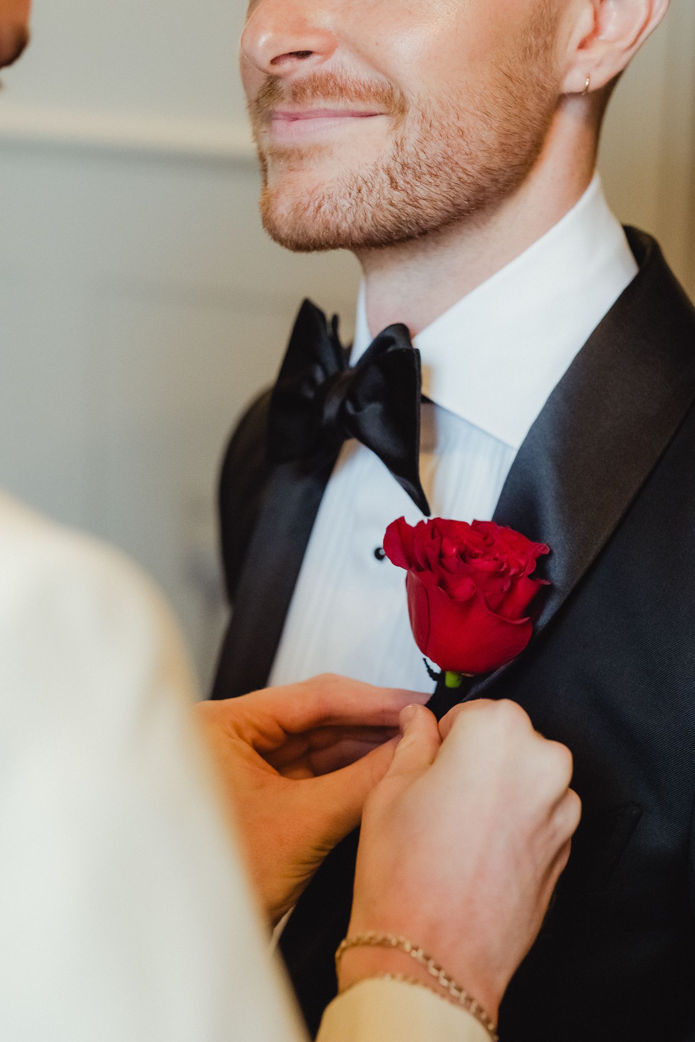 Close-up of a person in a black suit adjusting a vibrant red rose boutonniere on their lapel.