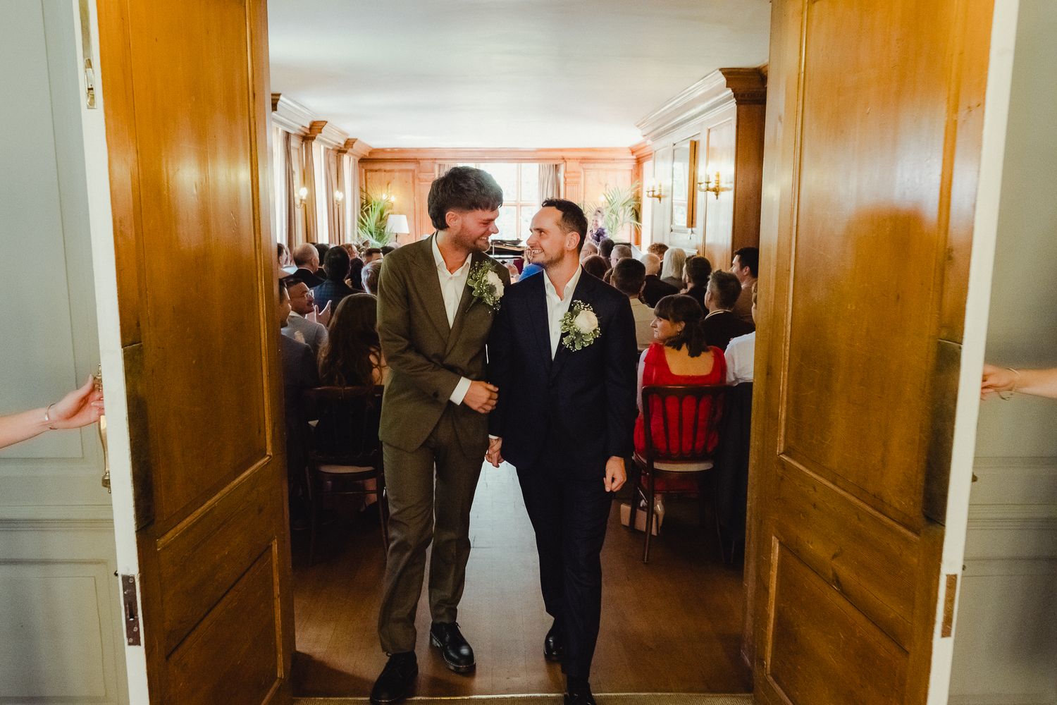 Two people in formal attire walking together through an elegant wood-paneled hallway filled with celebration guests.