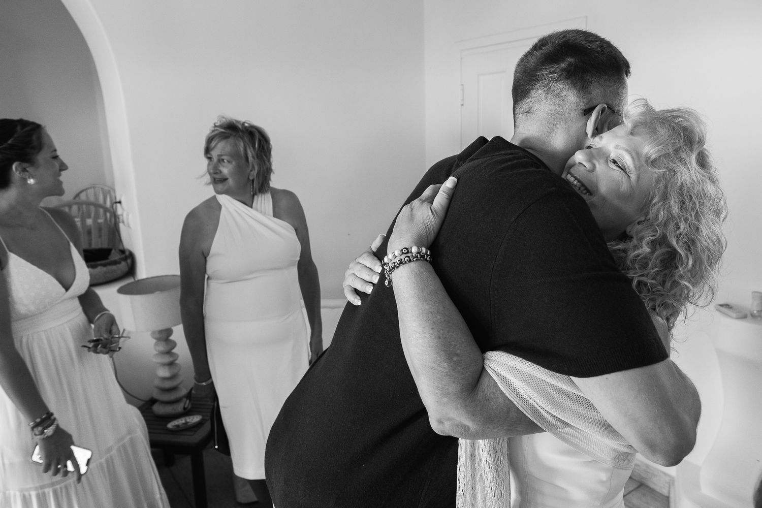 Black and white wedding photo of couple embracing warmly while guests smile nearby.