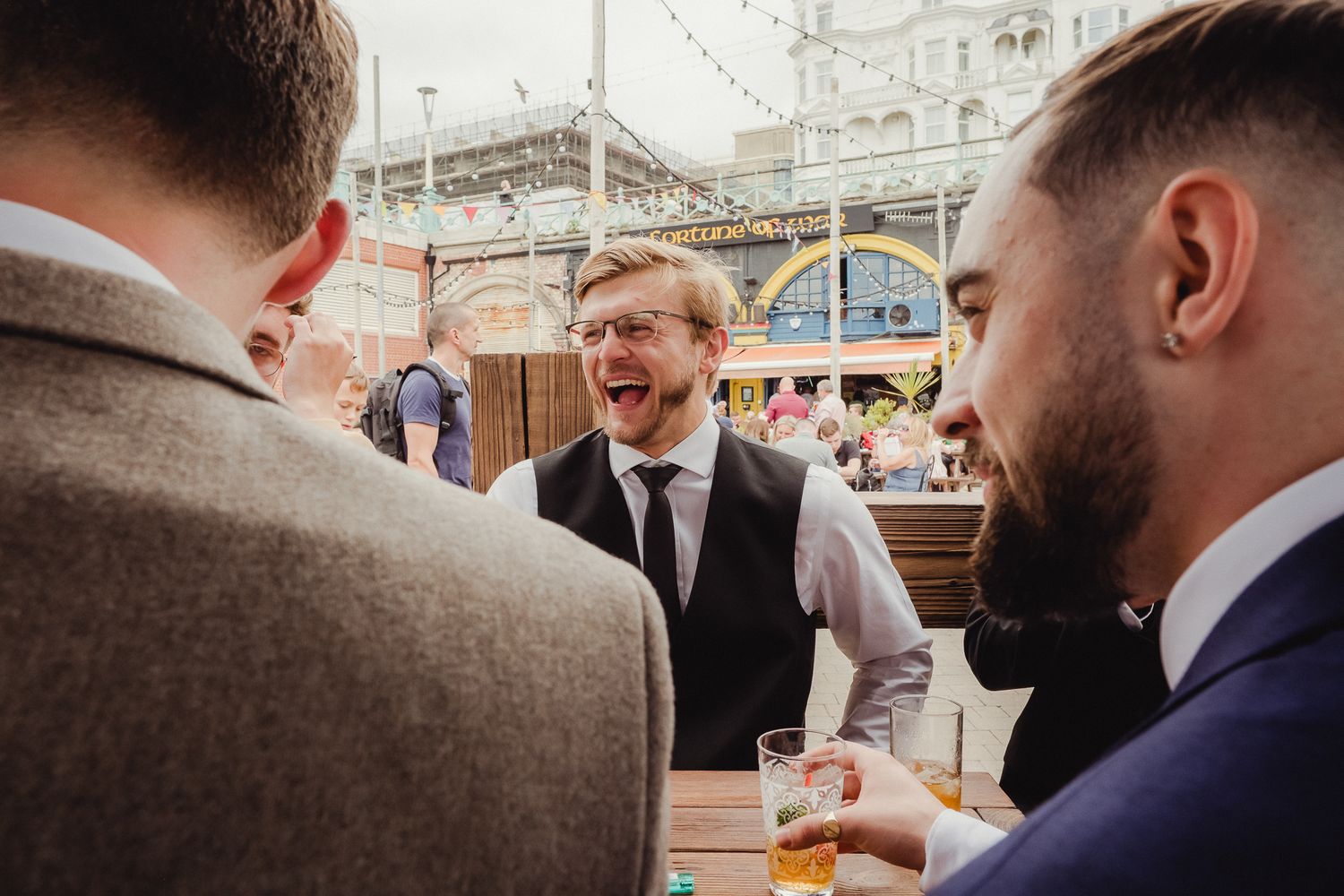 Groomsmen share a laugh and drinks at an outdoor wedding reception venue with decorative architecture in the background.