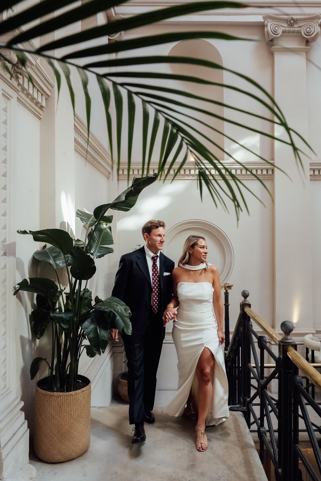 Couple in elegant wedding attire walking hand in hand through a bright, neoclassical interior with tropical palm leaves and potted plants.