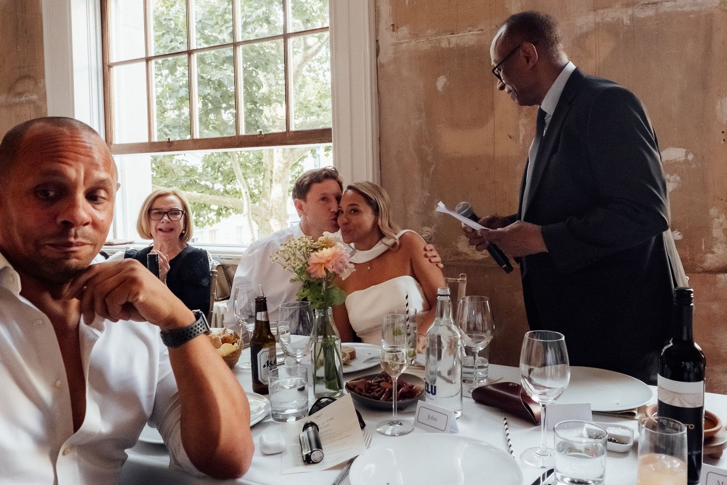 A man in a suit reads from a paper beside a smiling couple seated at a wedding table as another guest looks on.