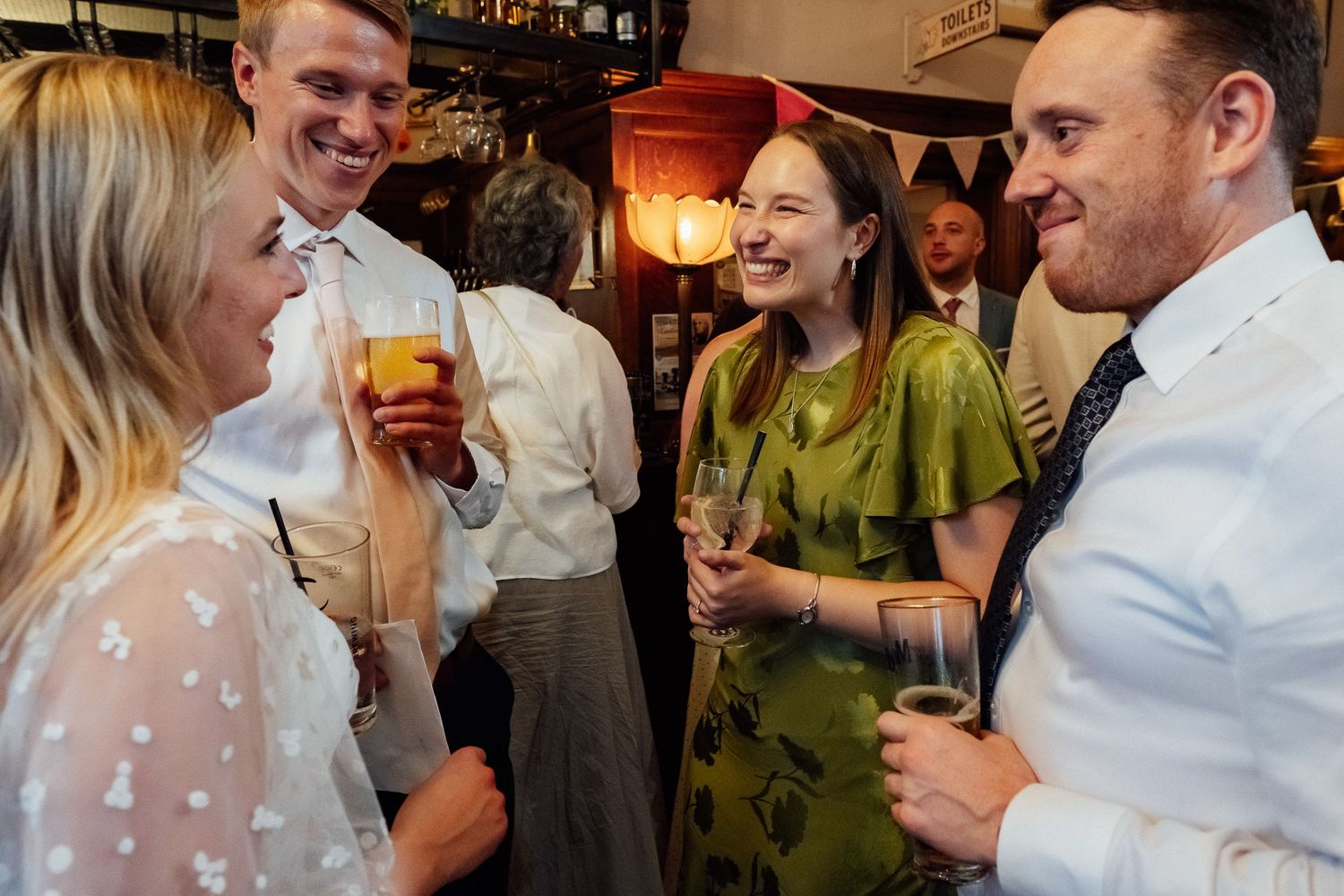 Smiling friends in smart casual attire hold drinks and chat at a lively pub gathering with warm lighting and bunting in the background.