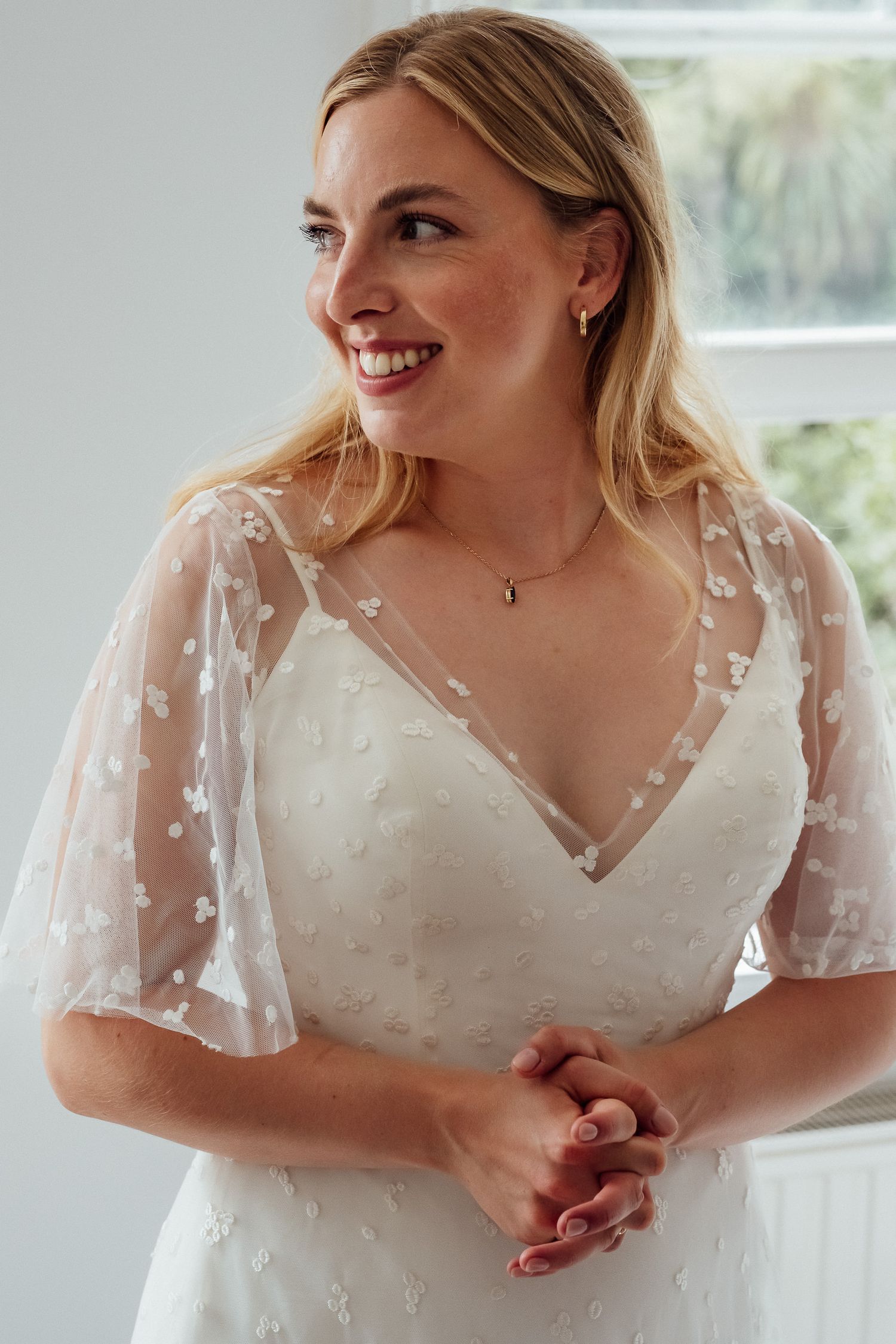 Smiling bride with long blonde hair wears a V-neck wedding dress with sheer floral flutter sleeves, hands clasped, looking to the side.