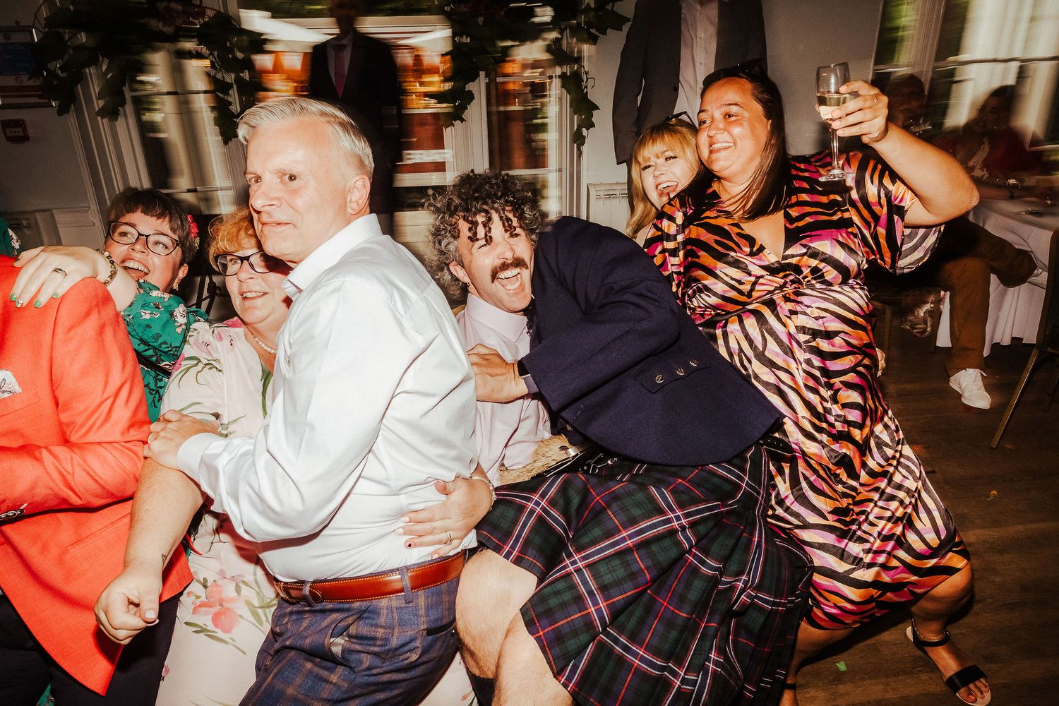 Group of joyful wedding guests laughing and dancing together, one man lifted in the air, at a lively indoor reception party.