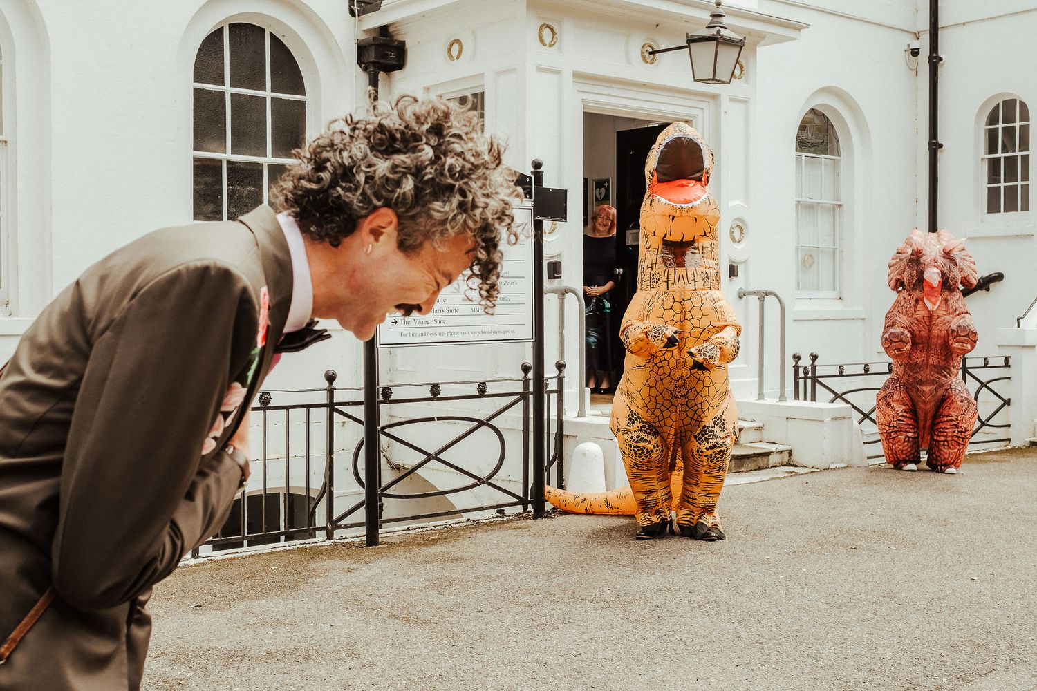 Curly-haired man in a brown suit bends forward laughing near two people in inflatable dinosaur costumes outside a white building.