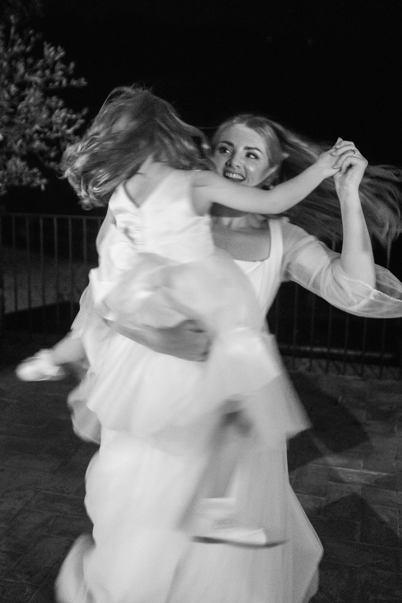 Black and white photo of a smiling woman in a white dress spinning joyfully while holding a little girl in a tulle dress outdoors at night.