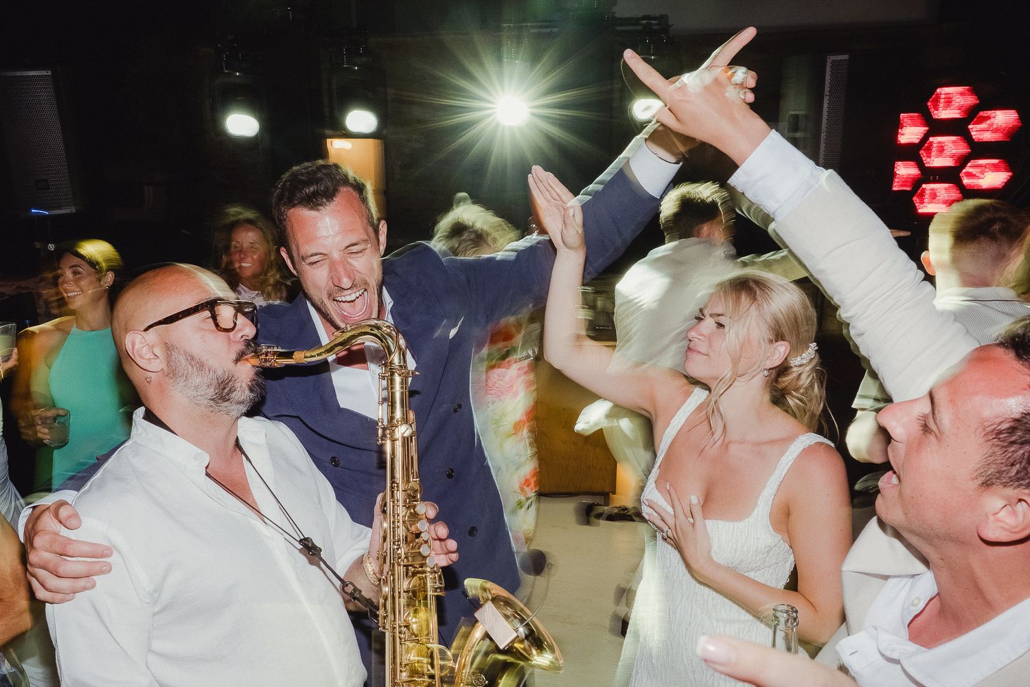 Energetic wedding party scene with live saxophone music and dancing guests in a festive indoor venue.