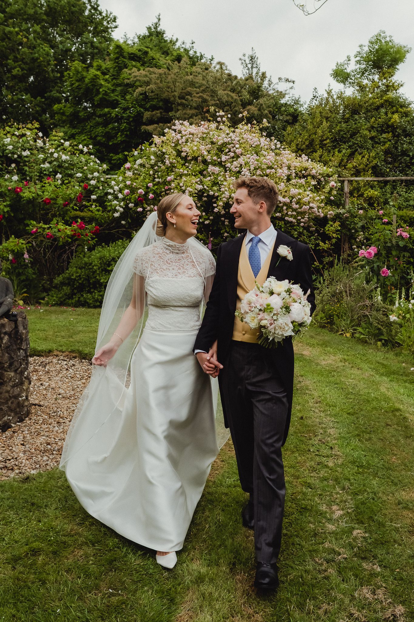 A couple on their wedding day walks together through a lush garden path surrounded by blooming flowers and greenery.