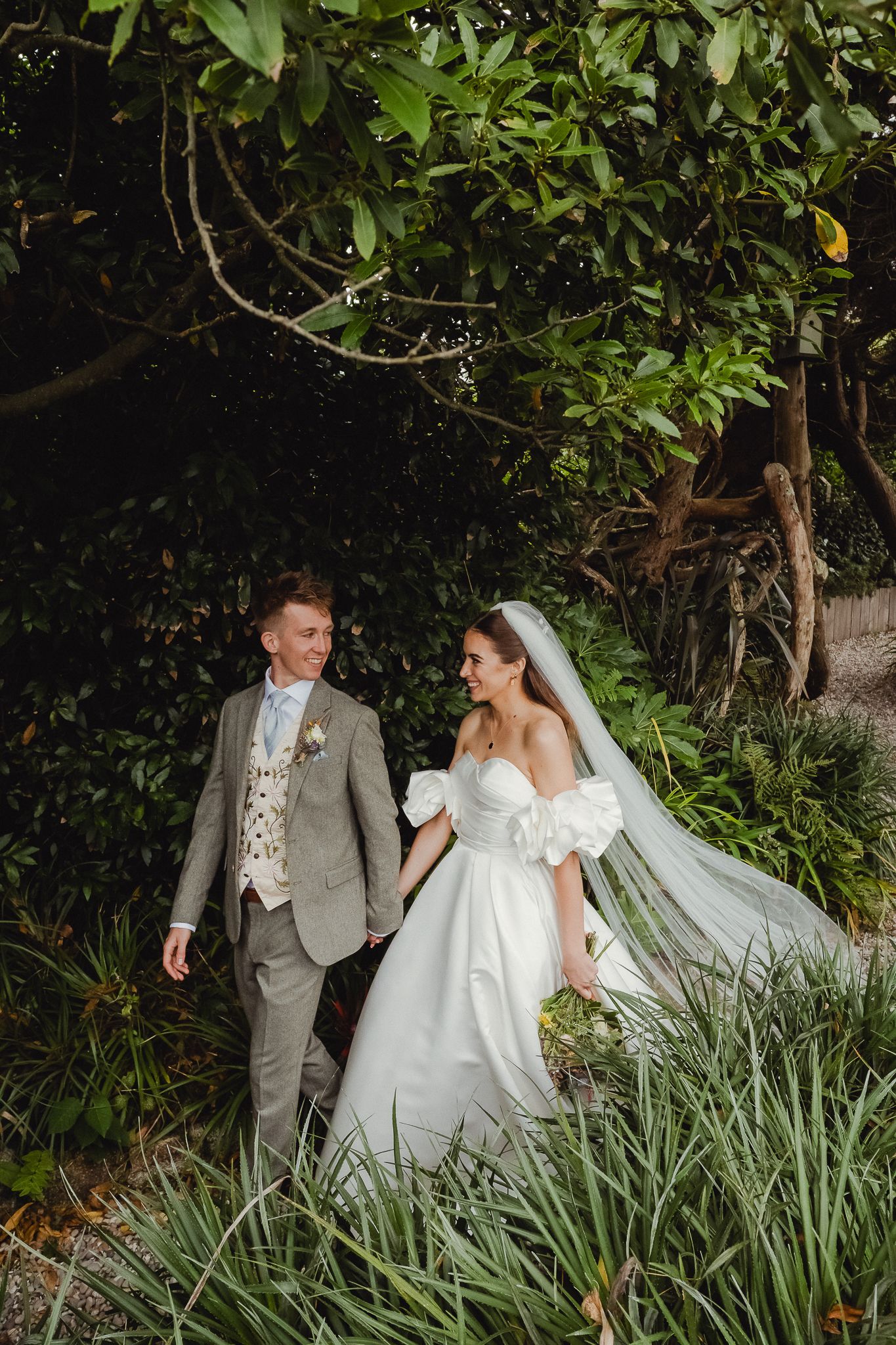 Bride and groom walk together through lush garden greenery during outdoor wedding photo session.