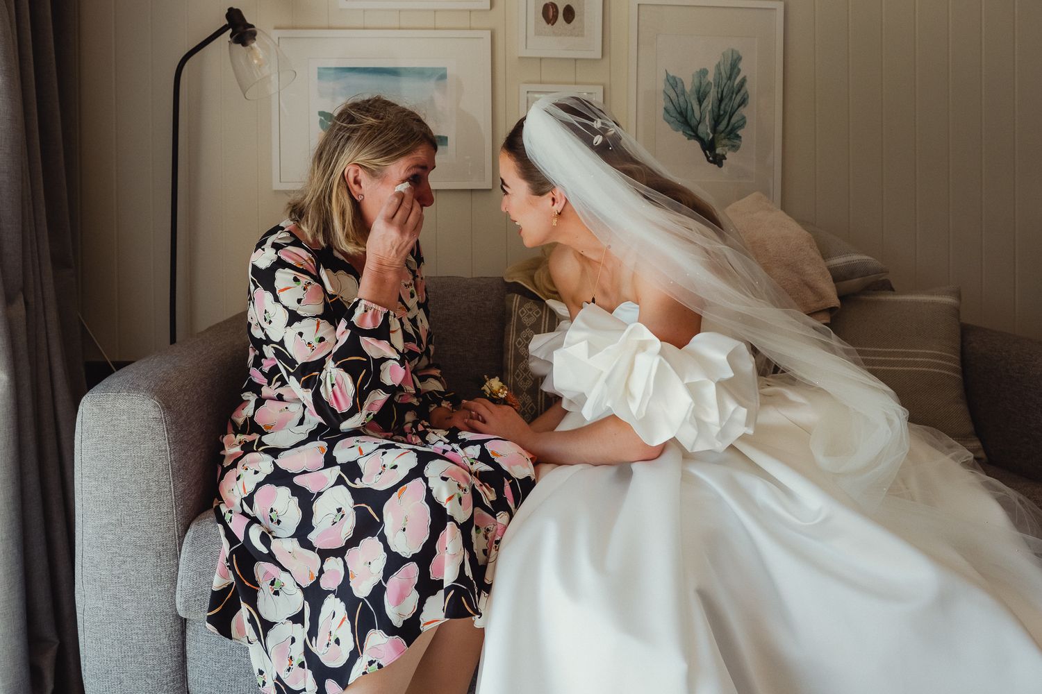 A person in a floral dress helps adjust a flowing white wedding gown in a brightly lit room.