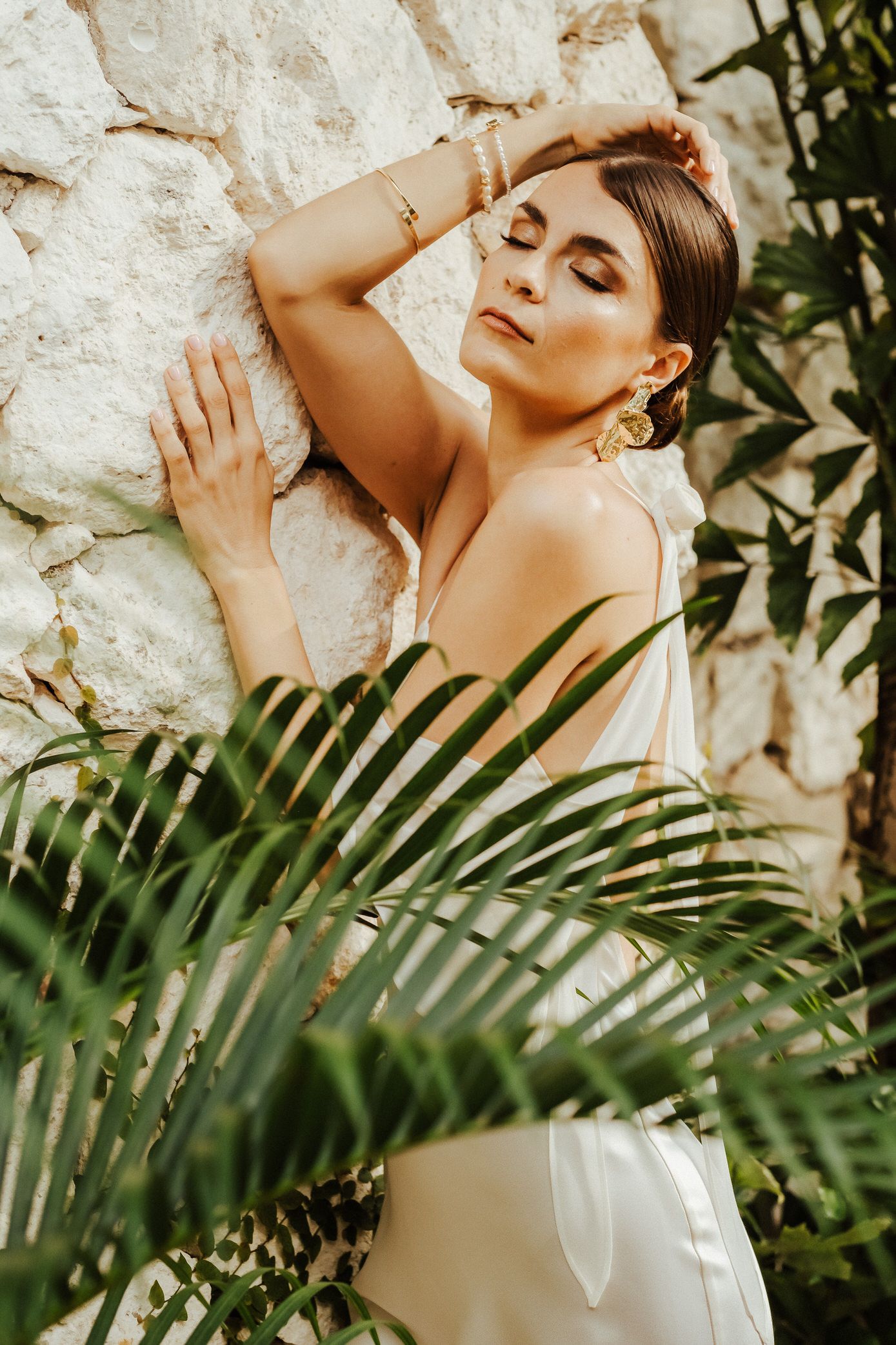 A fashion portrait of someone in a white dress posing against a stone wall with tropical palm leaves in the foreground.
