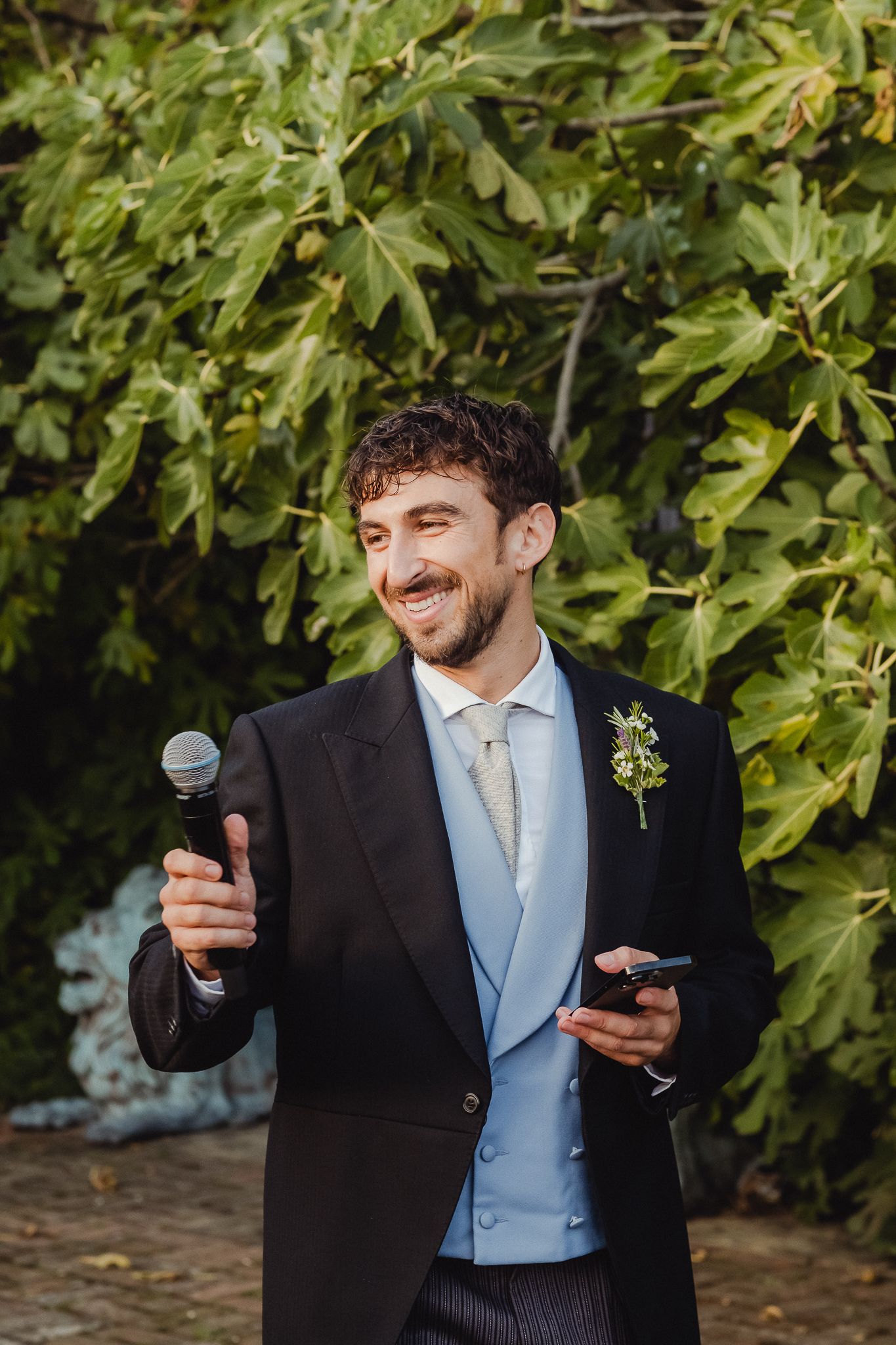 A groom in formal attire makes expressive gestures while holding a microphone during a wedding speech.
