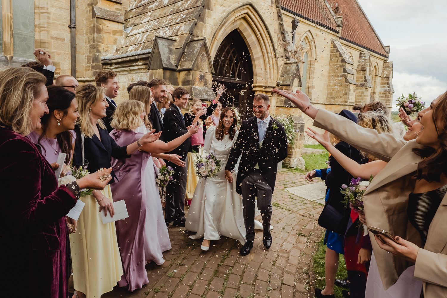 Wedding guests shower a couple with confetti as they exit a historic stone church with Gothic arched doorway.